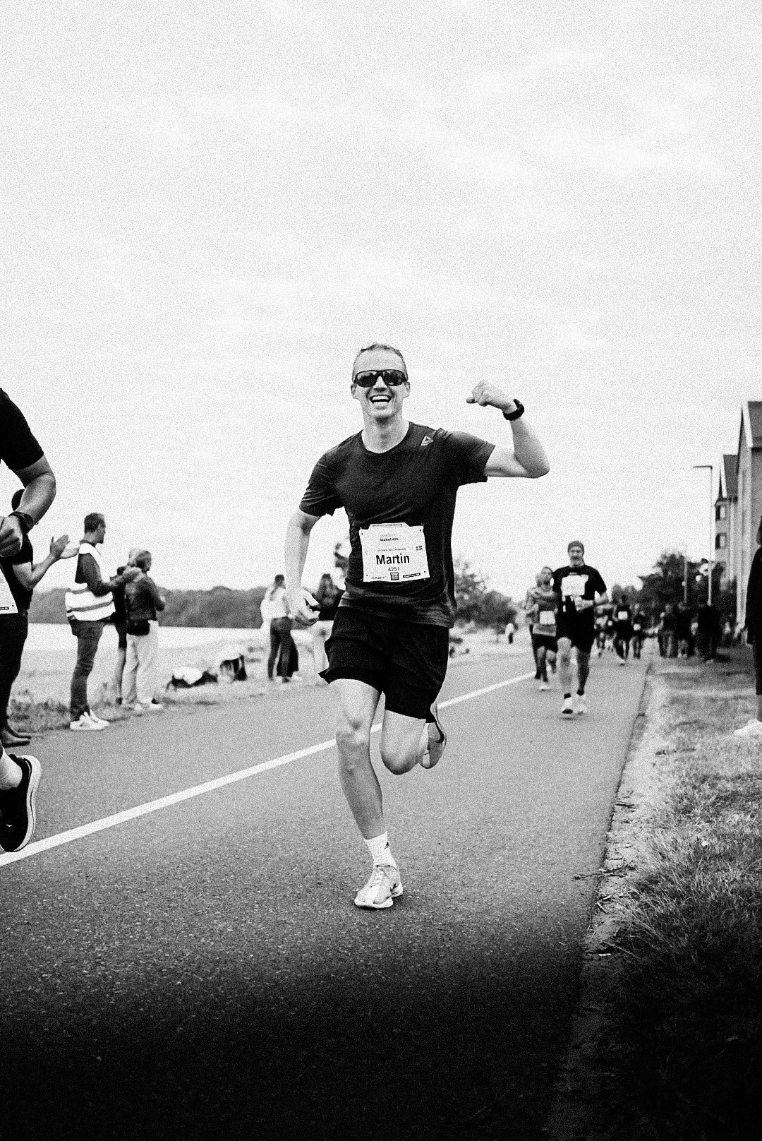 A woman running in a marathon, wearing sunglasses, a black shirt, and shorts, smiling and showing her bicep in a celebratory gesture. There are other runners and spectators along the roadside.