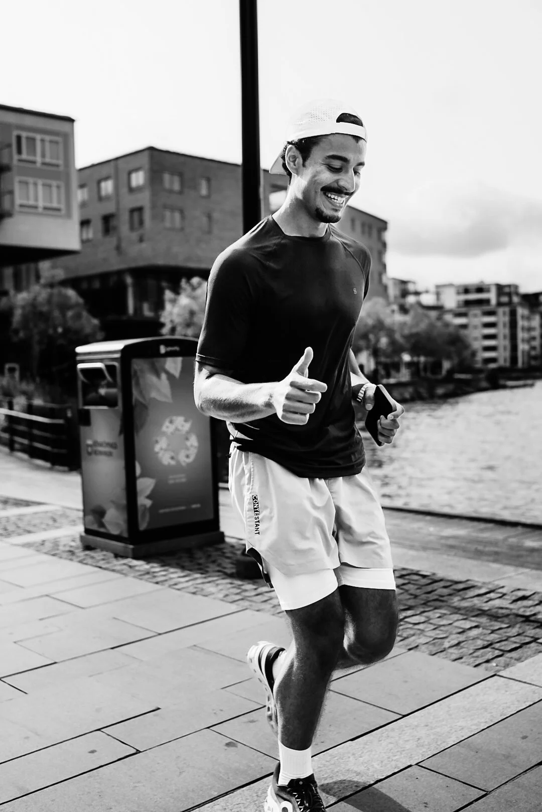 A young man running on a paved pathway near a body of water, smiling, dressed in a dark t-shirt, light shorts, and a backwards cap.
