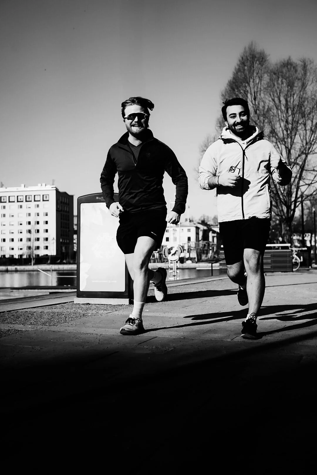 Two men jog along a city sidewalk during the day, with buildings and trees in the background, captured in black and white.