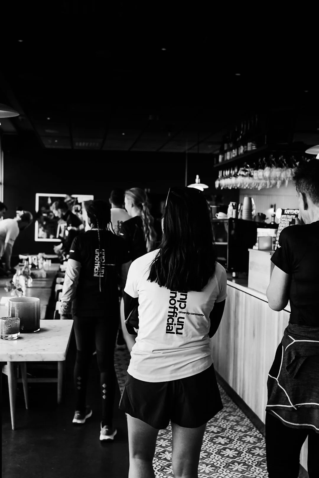 People standing in line inside a cafe or bar, some wearing uniforms or shirts with text on the back, with glasses hanging upside down above the counter.