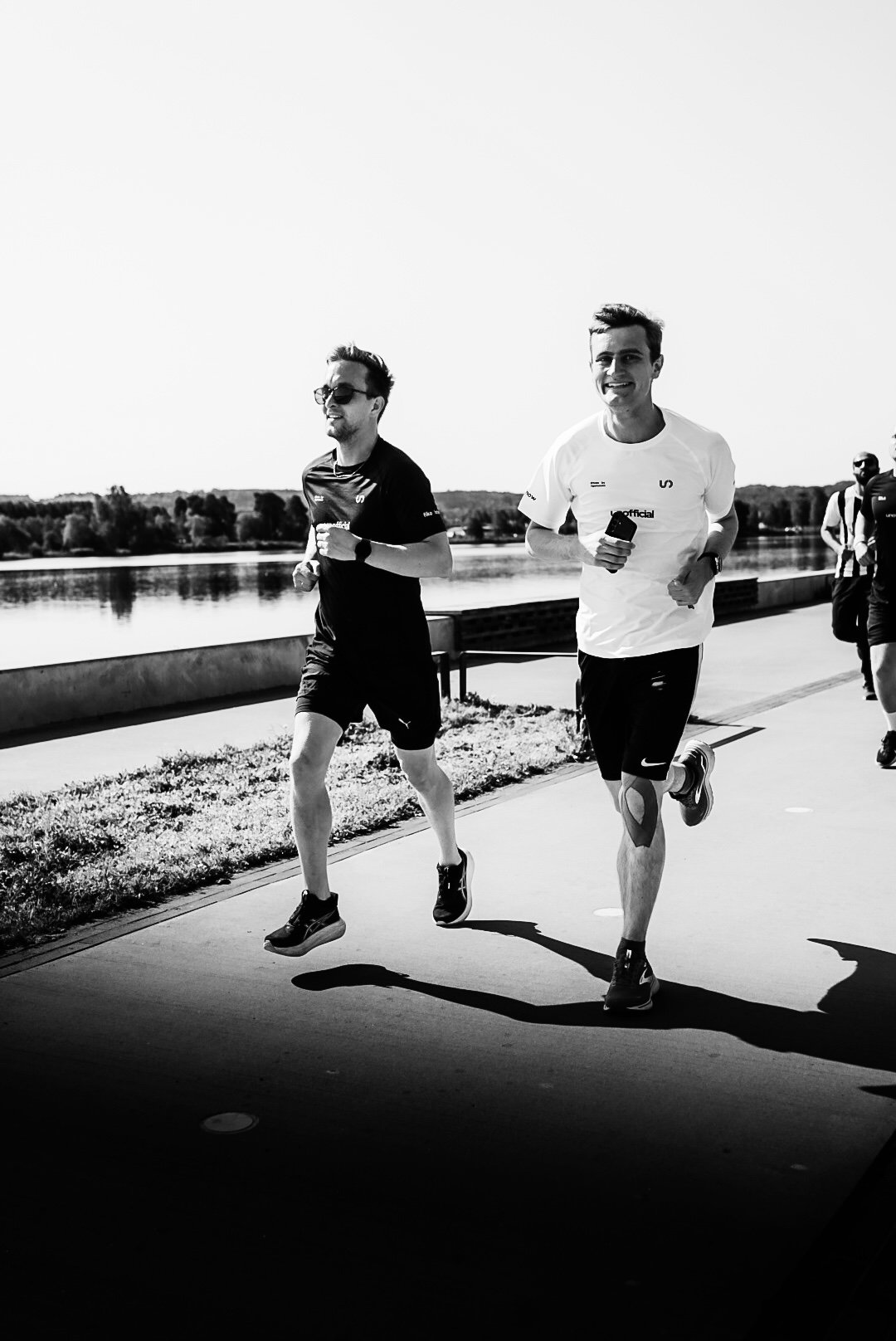Two men running along a riverside pathway during a race or marathon, with a scenic body of water and trees in the background.