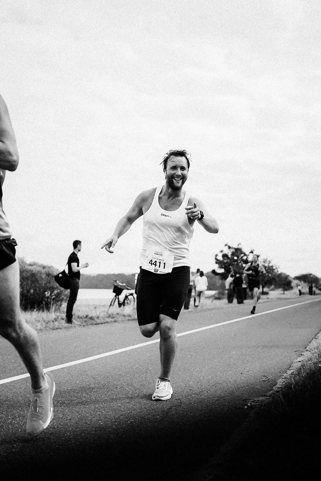 A man with a beard and short hair smiling. He wears a white tank top, black shorts, and running shoes, participating in a race near a body of water. Other runners and spectators are visible in the background.