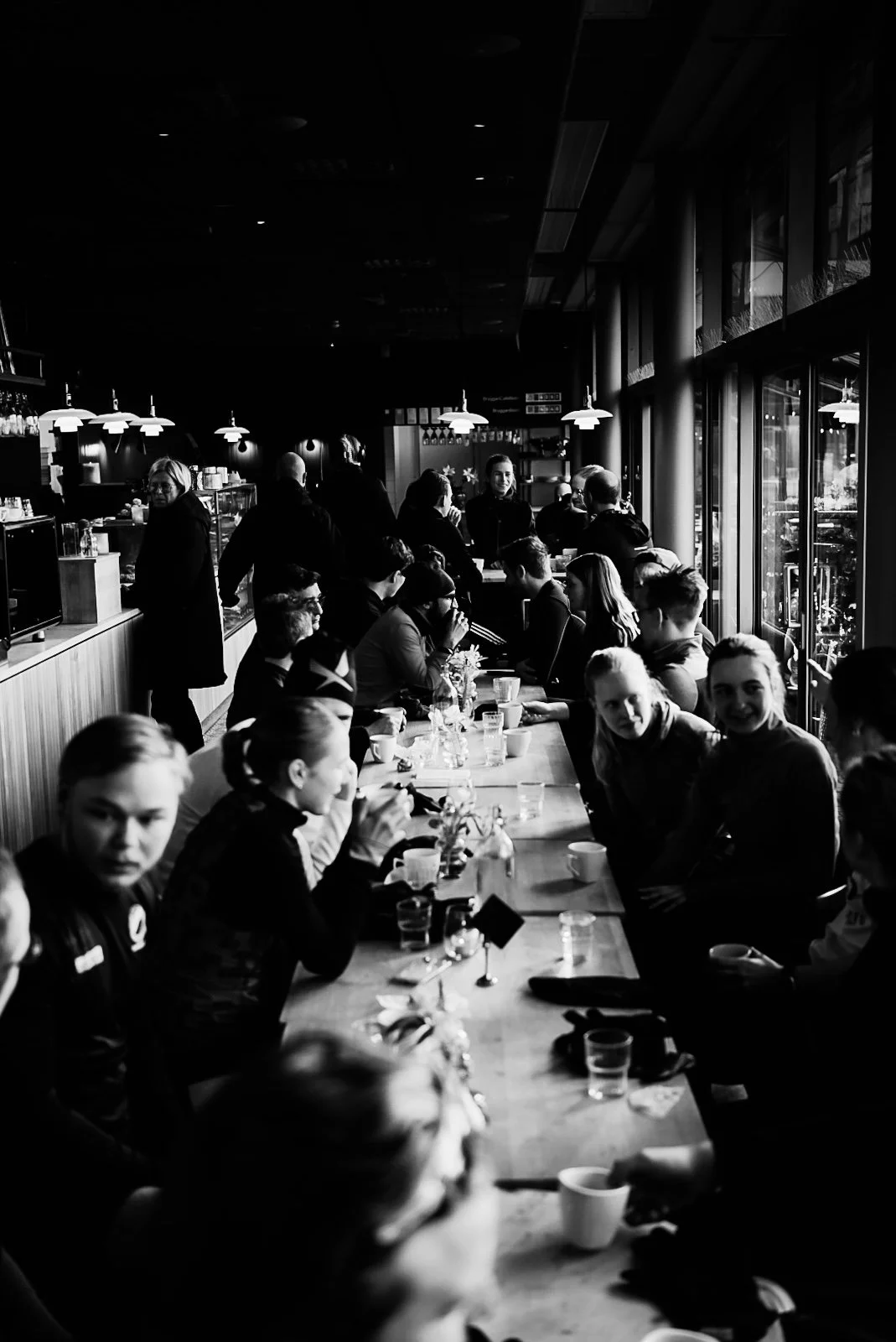 A group of people sitting at a long table in a cafe or restaurant, engaging in conversation, with some standing near the counter. The scene is in black and white, with a bright window on the right side of the image, afternoon sunlight shining in.