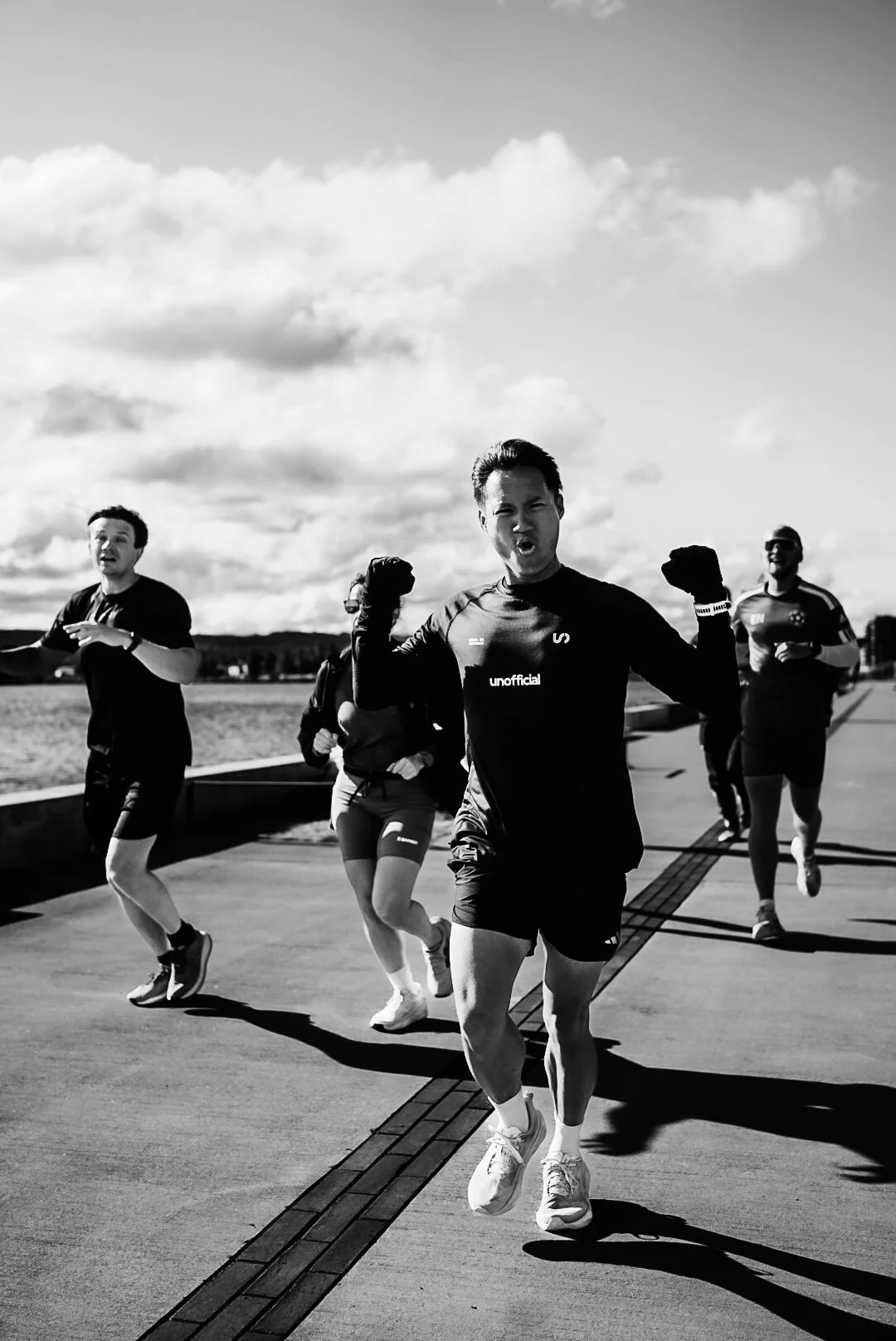 A group of five runners jogging outdoors on a paved path with a cloudy sky in the background. The runner in the front is a man wearing a black shirt and shorts, with a confident expression, flexing his arms.