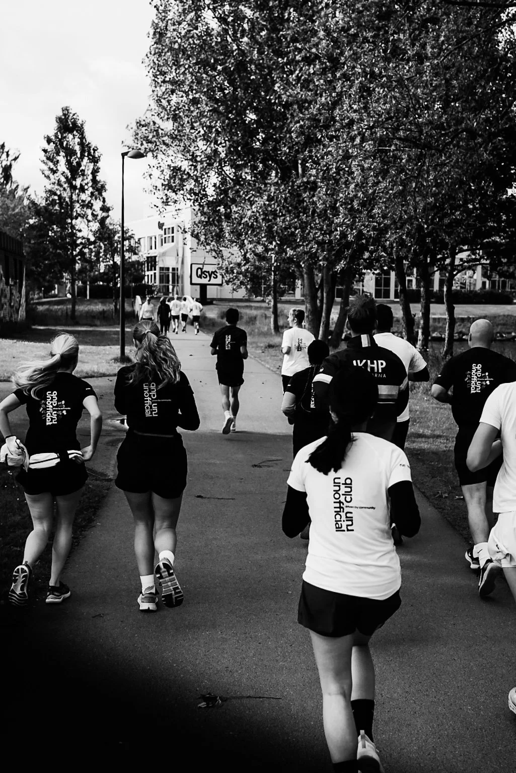 A group of people running outdoors on a paved path, wearing athletic clothes, with trees and buildings in the background.