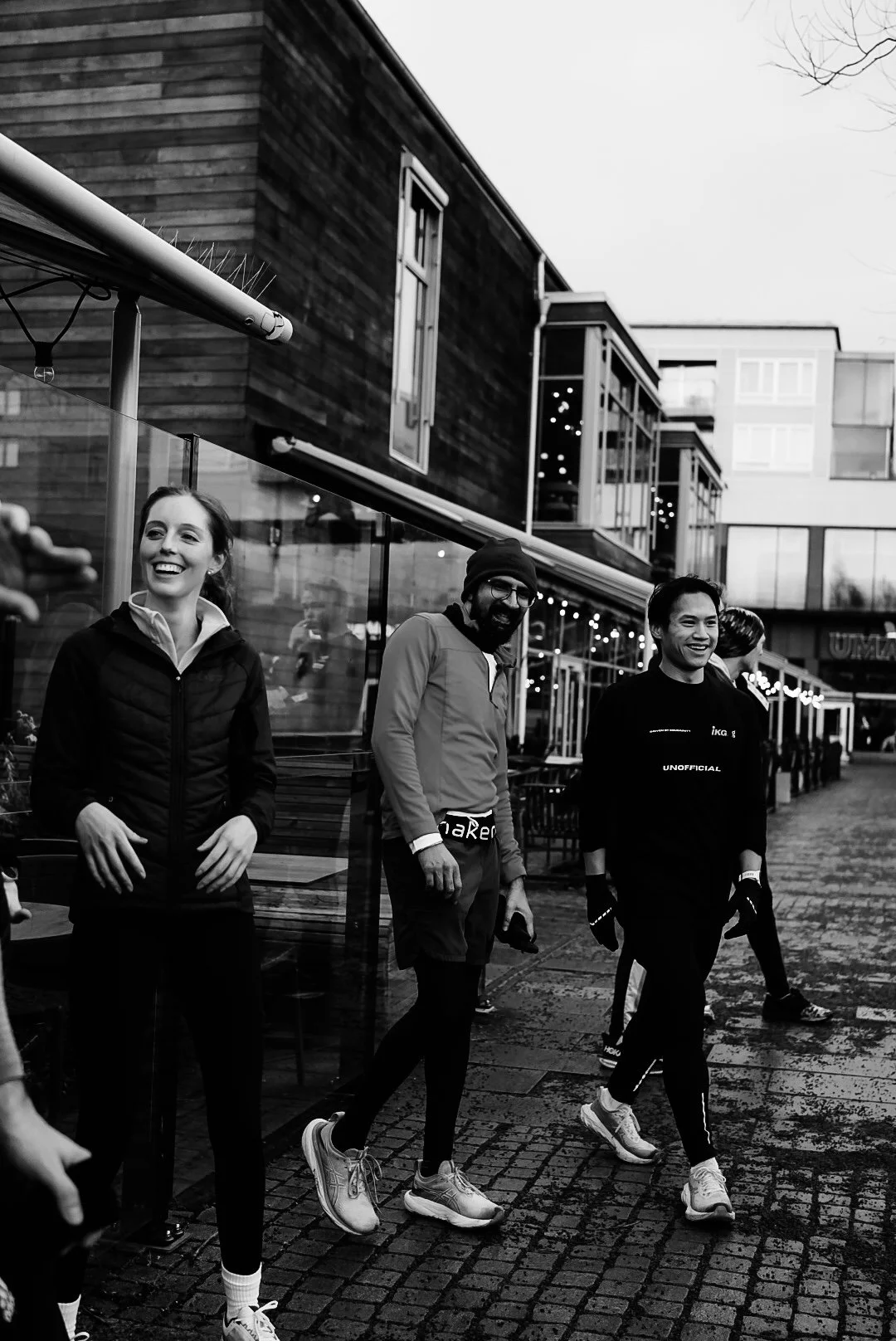 Three people outdoors smiling and talking, standing on a cobblestone sidewalk near a modern building with large windows and string lights.