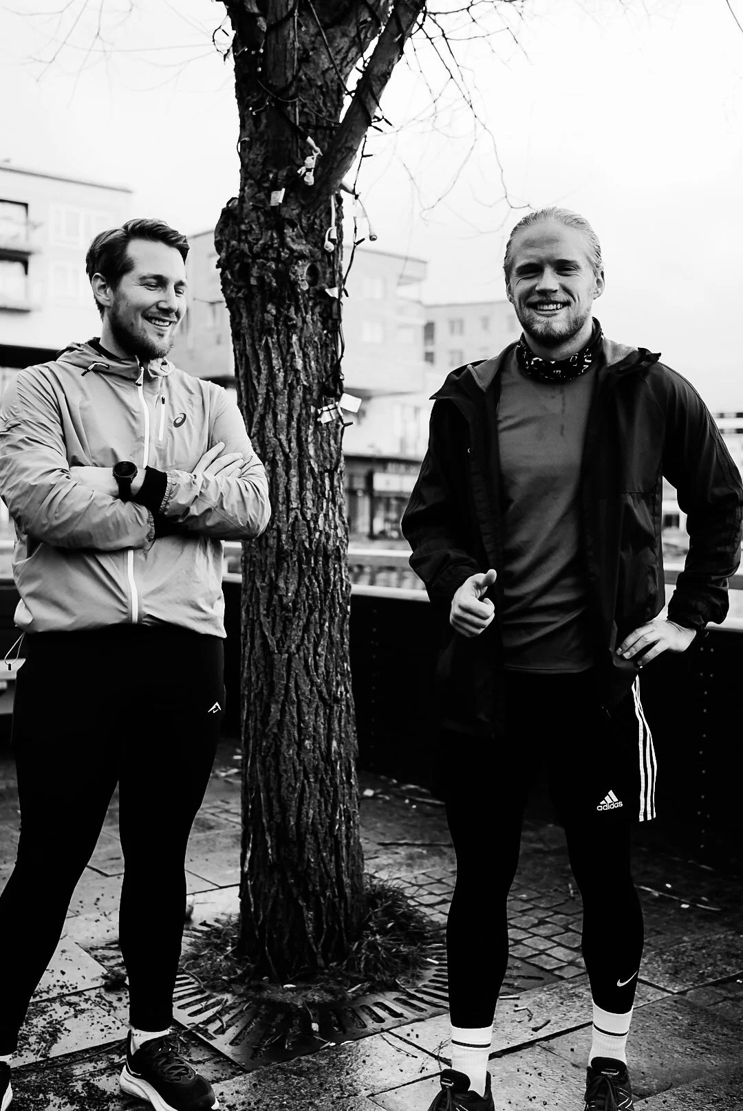 Two men standing outdoors near a tree, smiling. One has arms crossed, the other has hand on hip and is giving a thumbs-up. Both are dressed in athletic clothing.