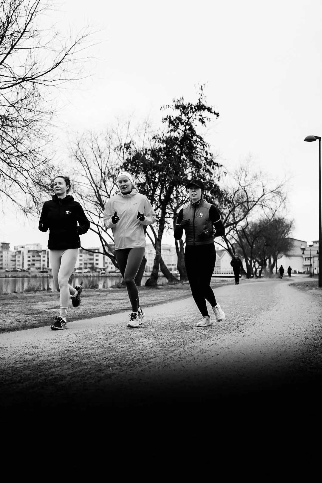 Three women running outdoors on a path with trees and buildings in the background in black and white.