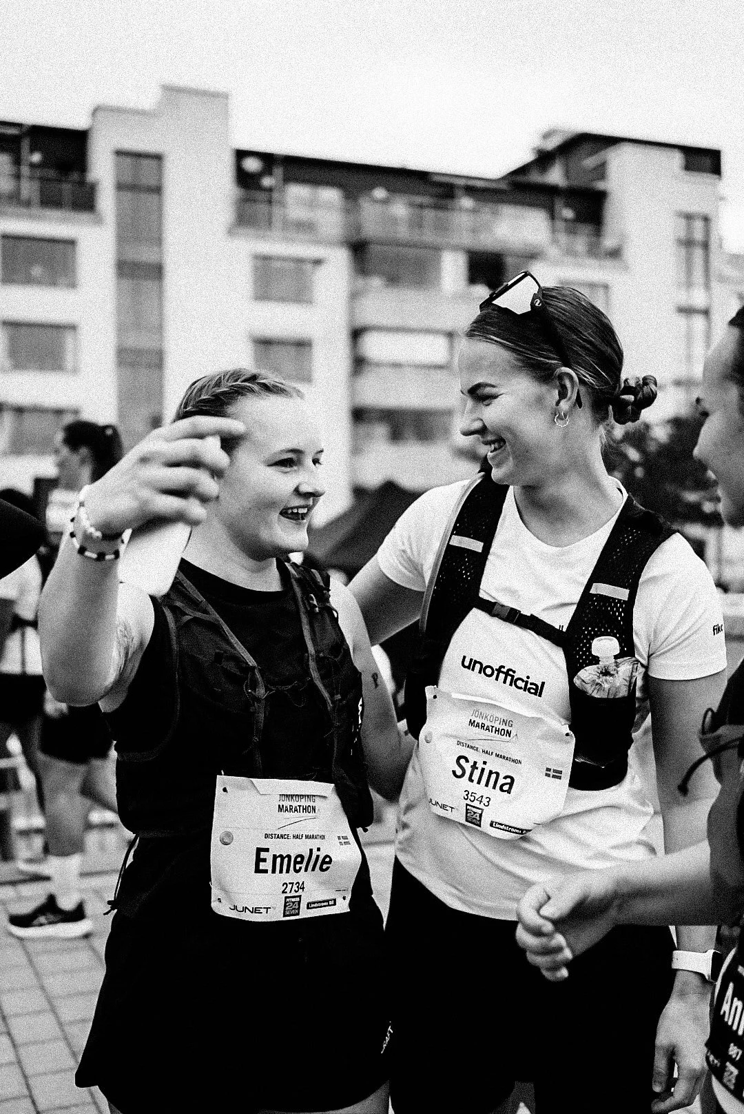 Two women smiling and talking during a marathon, with race bibs on their clothing, in an urban setting.