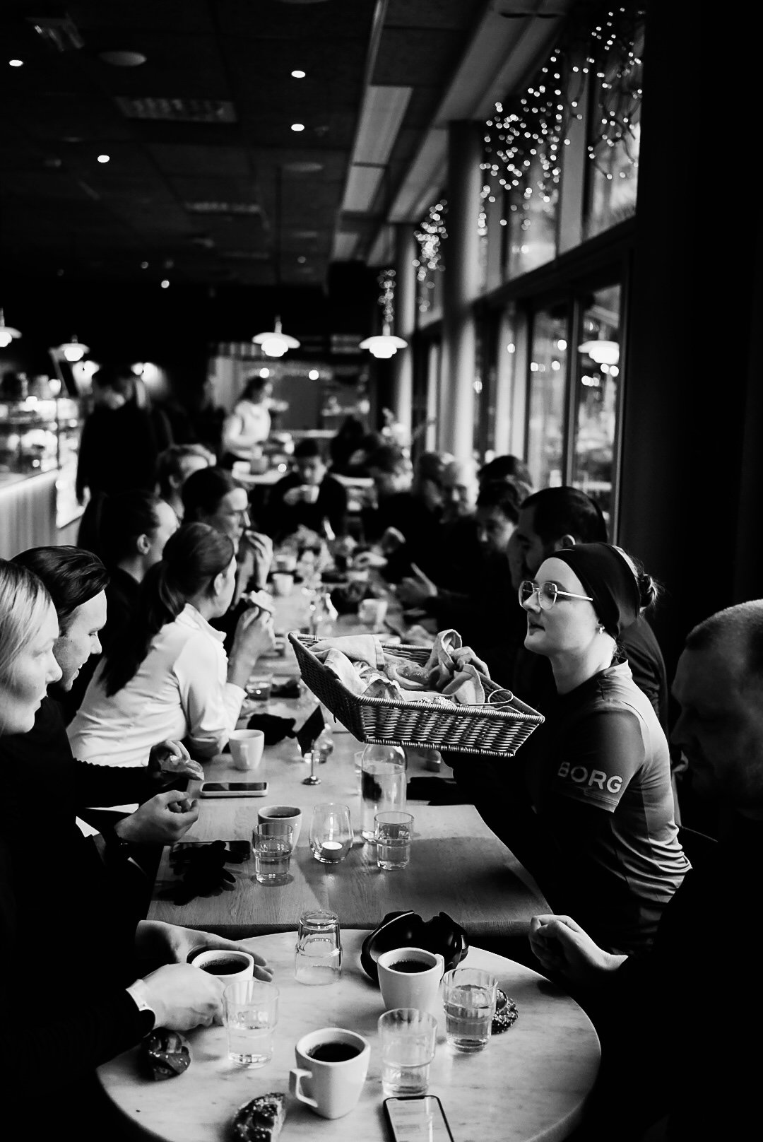 Black and white photo of a group of people sitting at a long table in a cafe or restaurant, some with cups of coffee, with a woman holding a basket of baked goods or food. Large windows with string lights are visible on the right side.