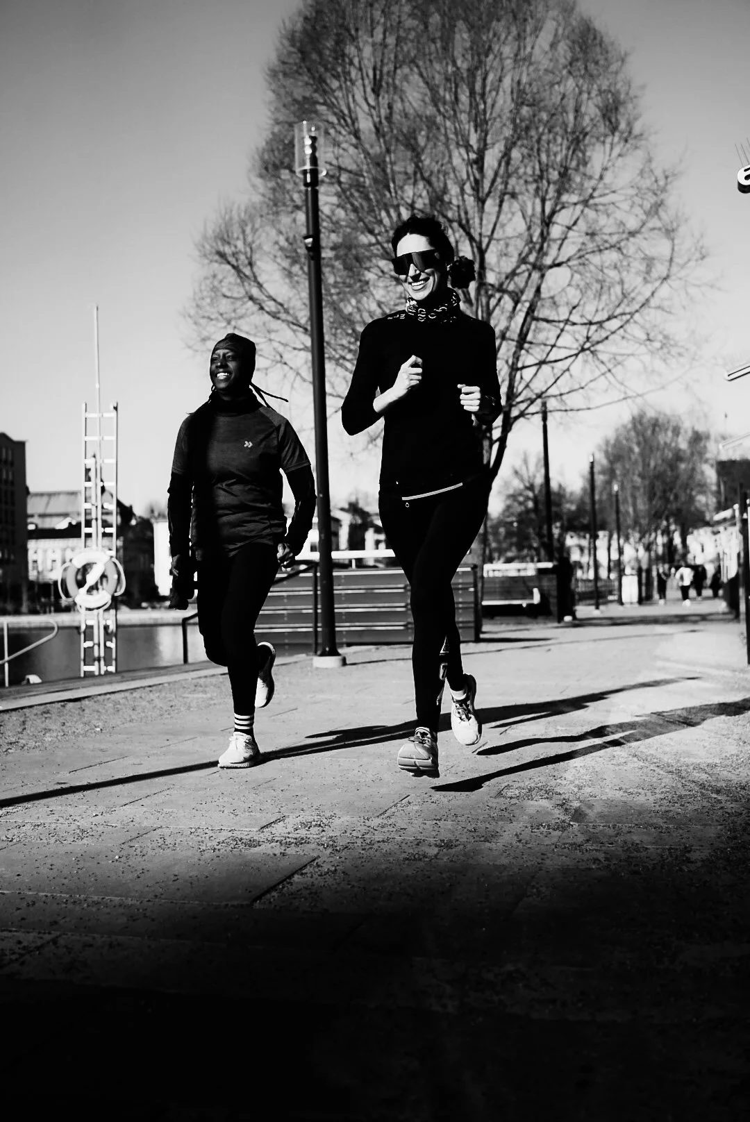 Black and white photo of two women jogging outdoors on a sunny day, with leafless trees and a clear sky in the background.
