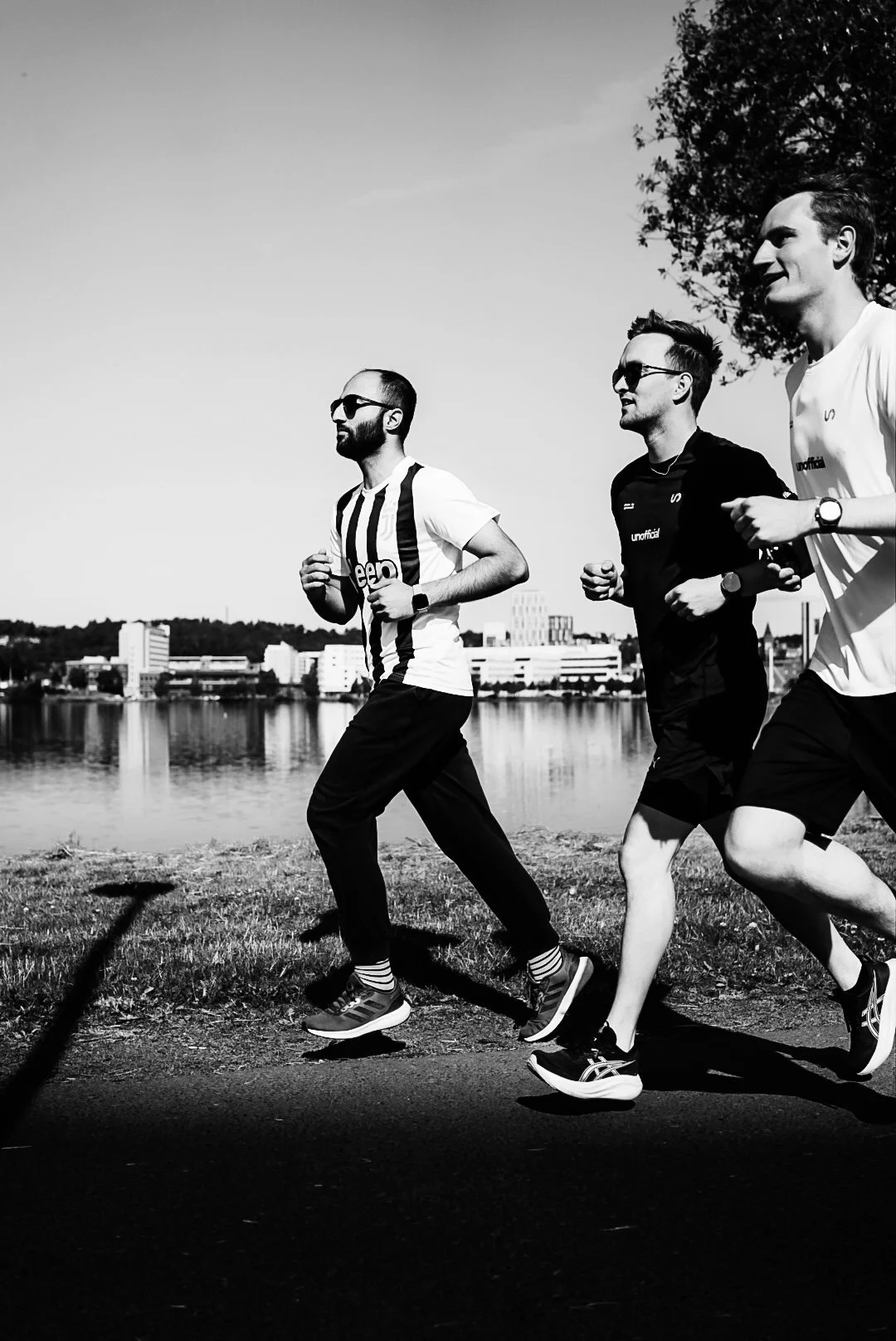 Three men running outdoors near a body of water and city buildings in the background, black and white photo.