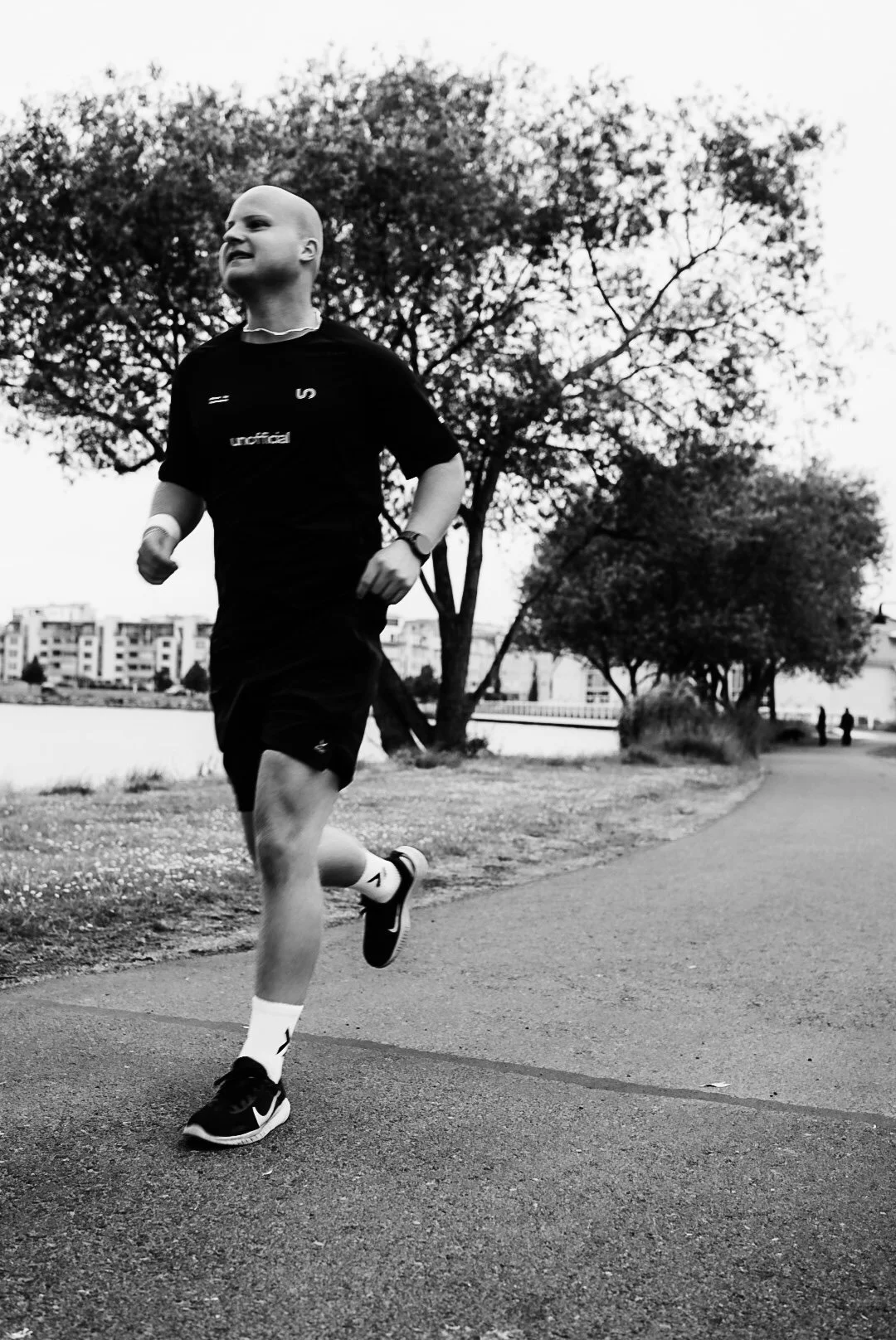 A young person running on a sidewalk near a body of water, with trees and buildings in the background, in black and white.