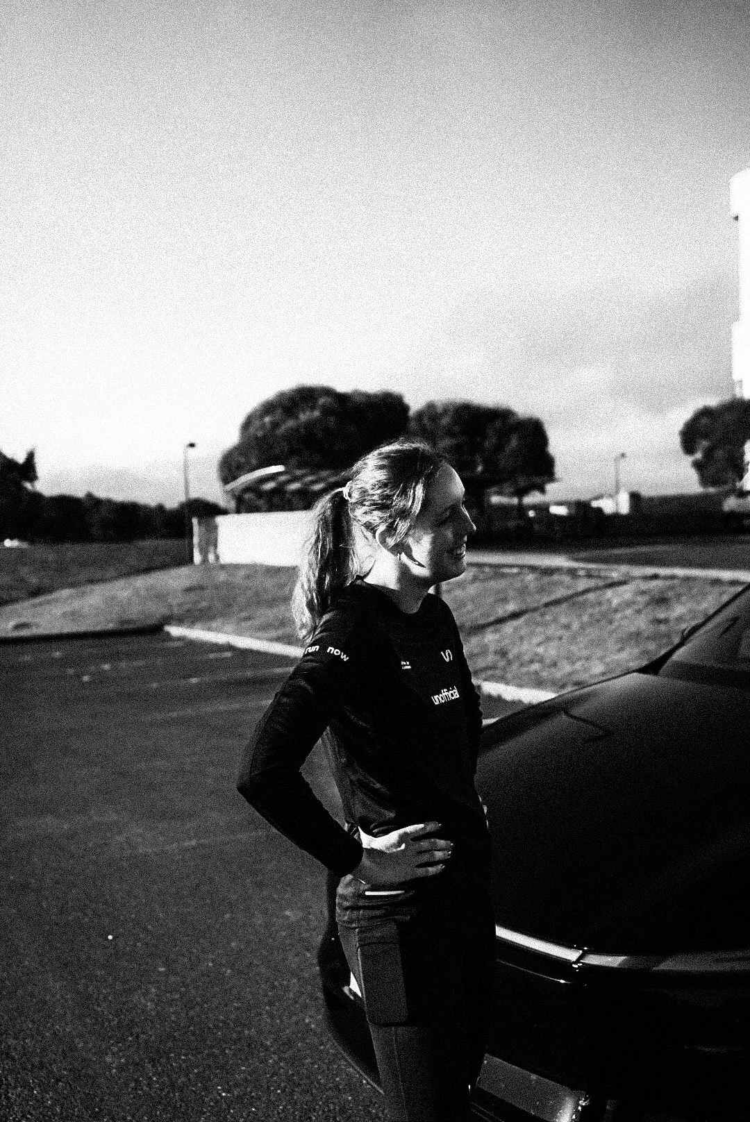 A woman with a ponytail stands beside a car in a parking lot, smiling, with trees and streetlights in the background during dusk.