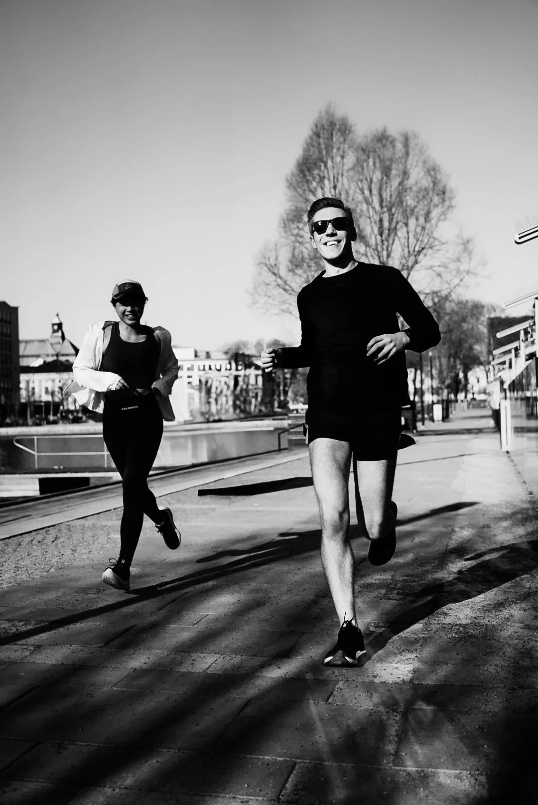 Two people running outdoors on a sunny day, smiling, with trees and buildings in the background. In black and white.