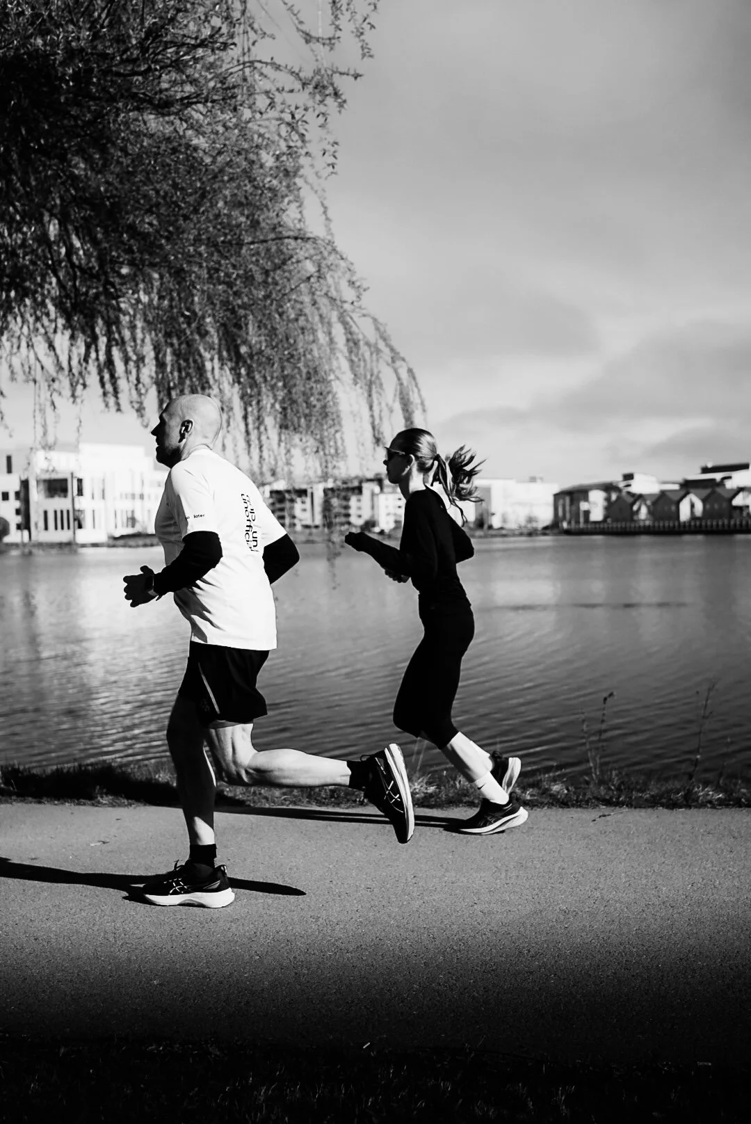 Black and white photo of a man and woman jogging along a waterfront path, with buildings and trees in the background.