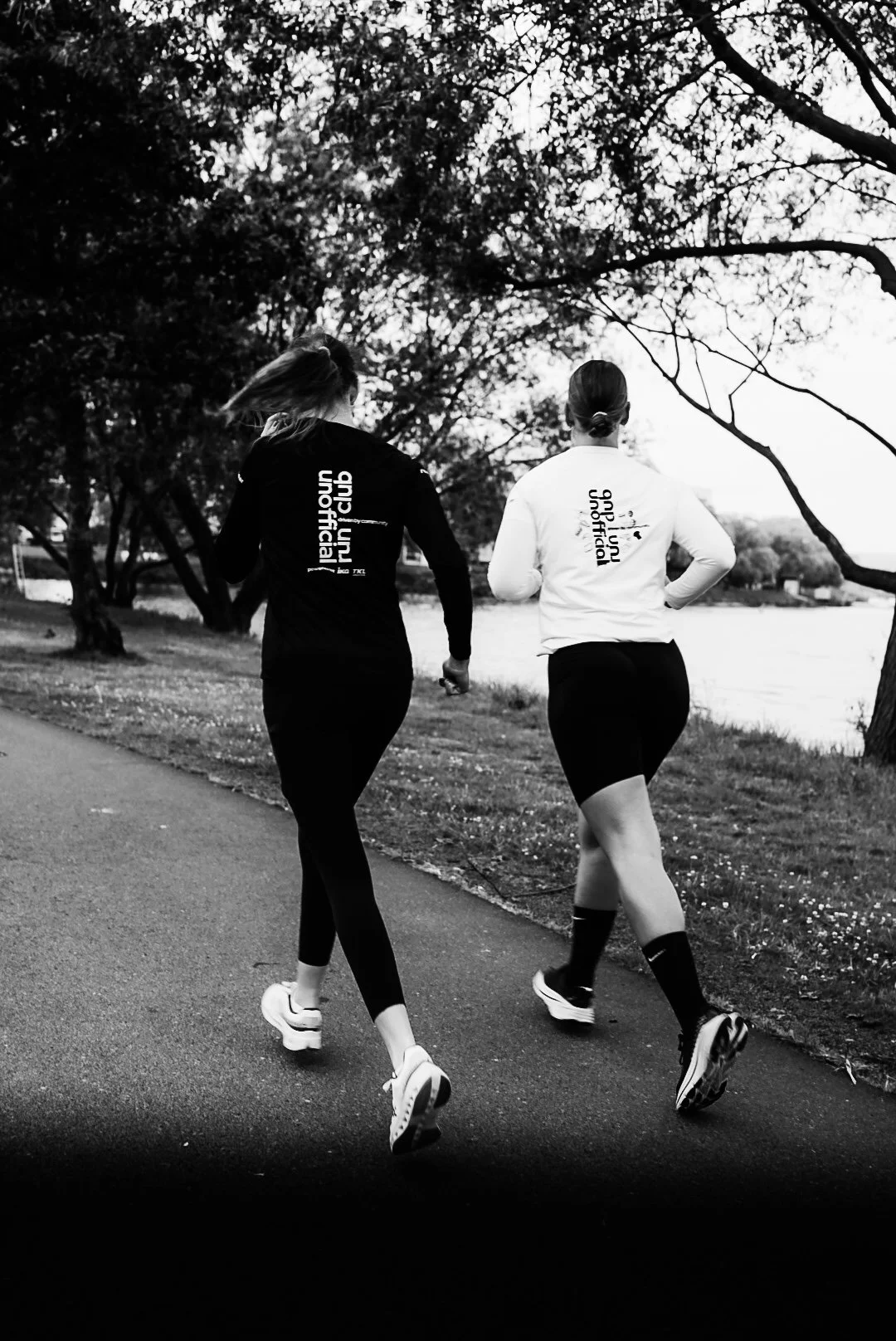 Two women jogging outdoors near a river, with trees and a cloudy sky in the background.