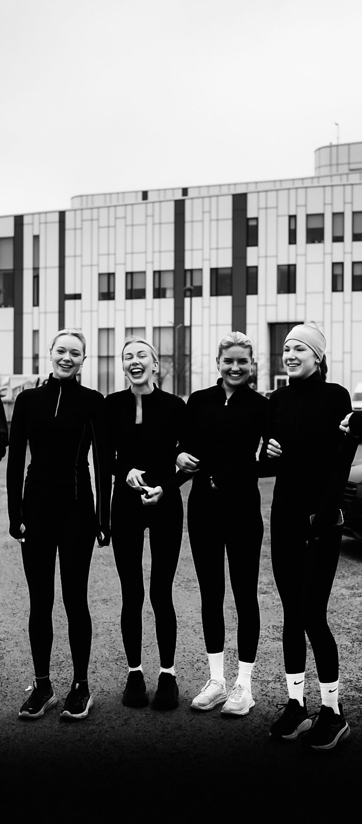 Four women in athletic clothing standing outside, smiling, in front of a modern building.