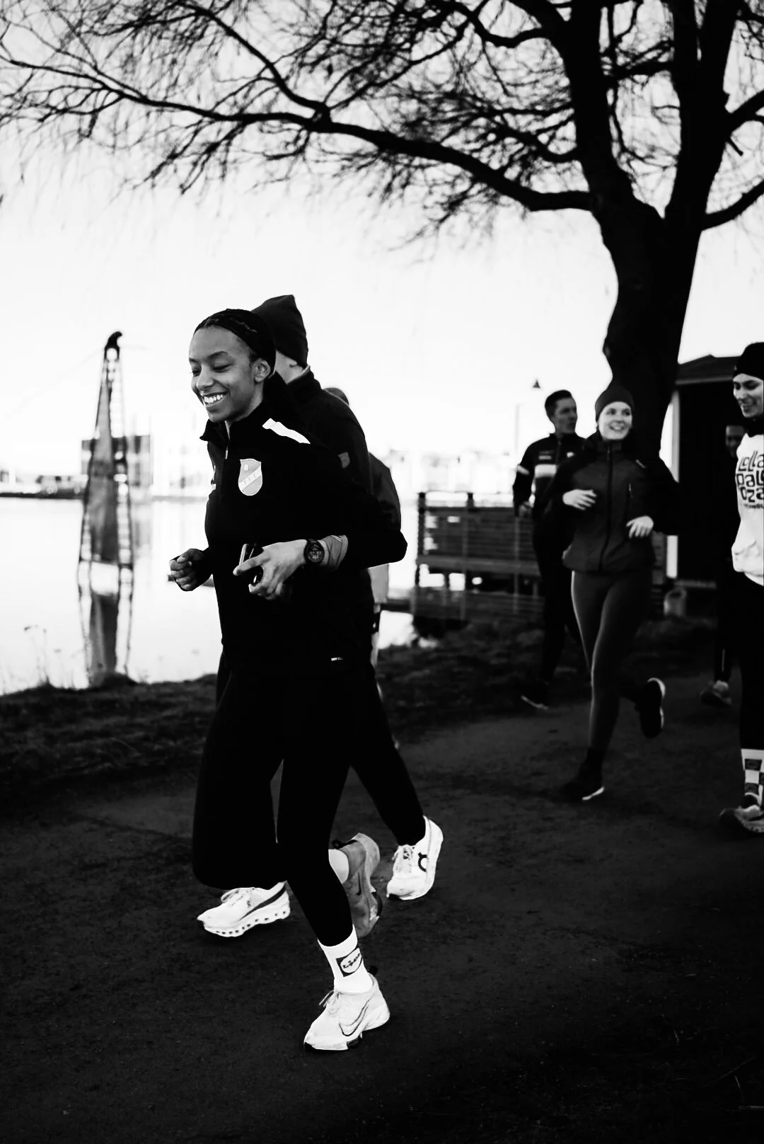 A group of people jogging outdoors near a body of water, smiling and dressed in athletic wear, with trees and a dock in the background.