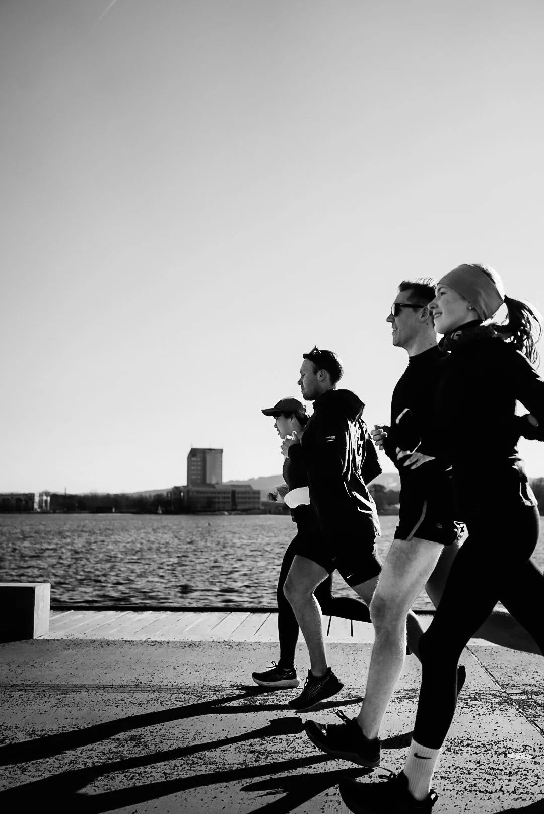 Four people running along a waterfront during sunrise or sunset, with a cityscape in the background, depicted in black and white.