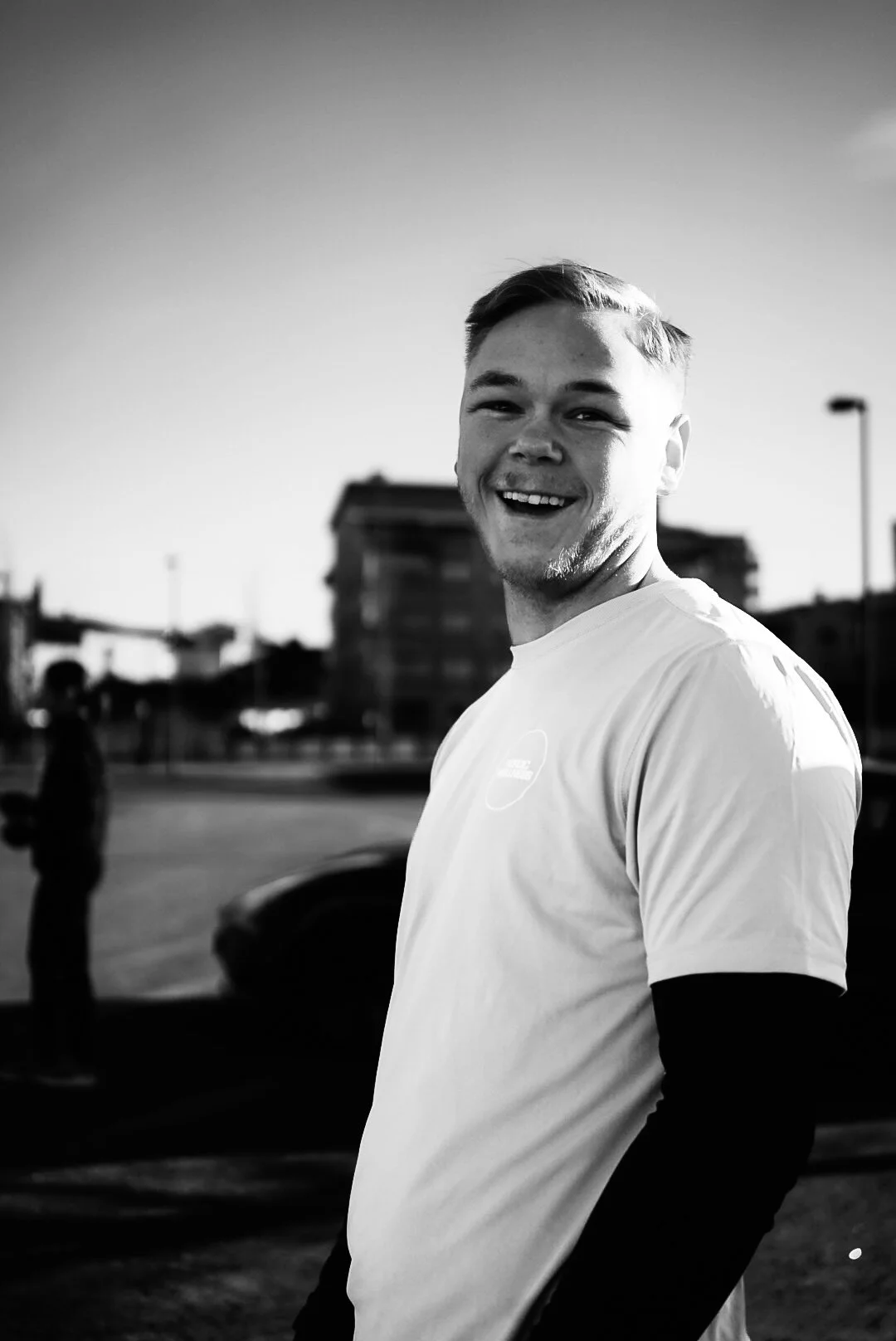 A young man standing outdoors, smiling, with buildings and a person in the background, captured in black and white.