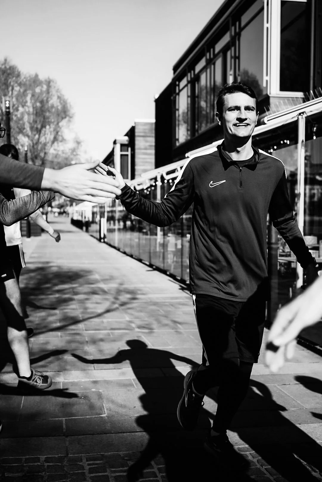 A man in athletic clothing running outside while giving a high five to someone off-camera during daytime.