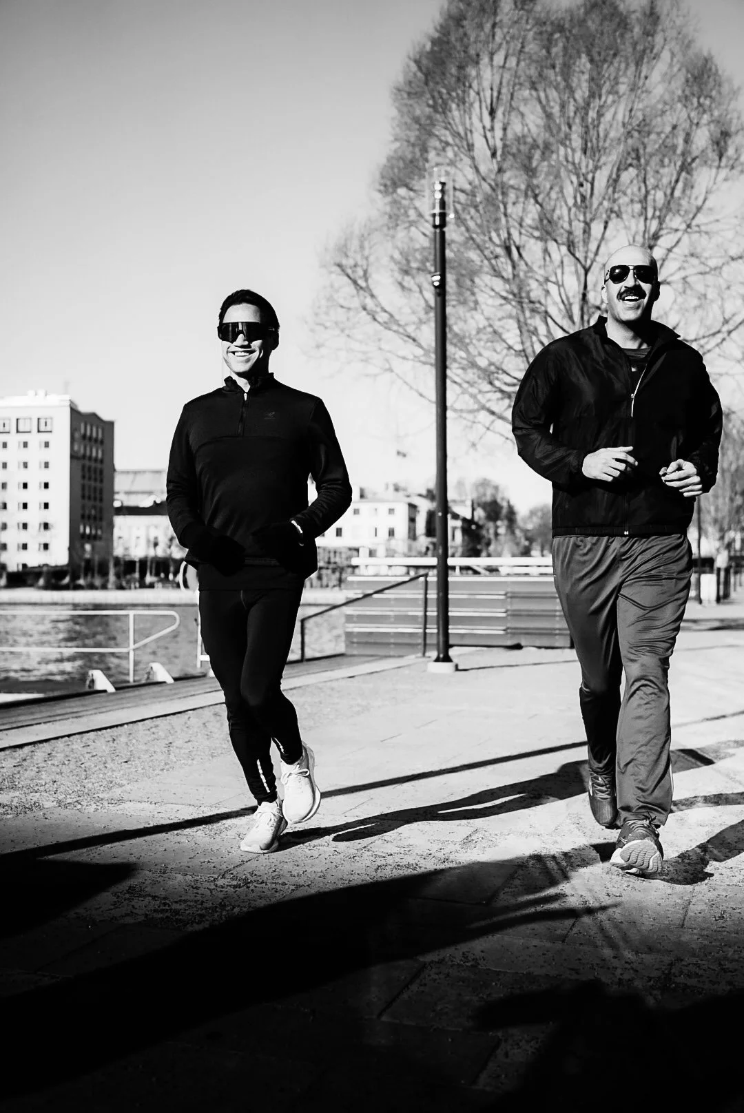 Two men jogging outdoors during daytime, wearing sunglasses and athletic clothing, with trees and buildings in the background.