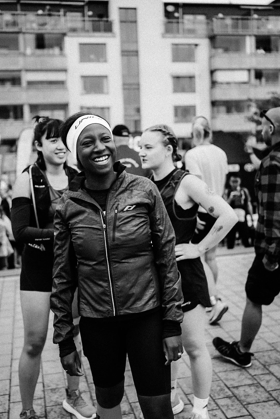 Smiling woman in athletic gear at a running event, surrounded by other runners and spectators outdoors.