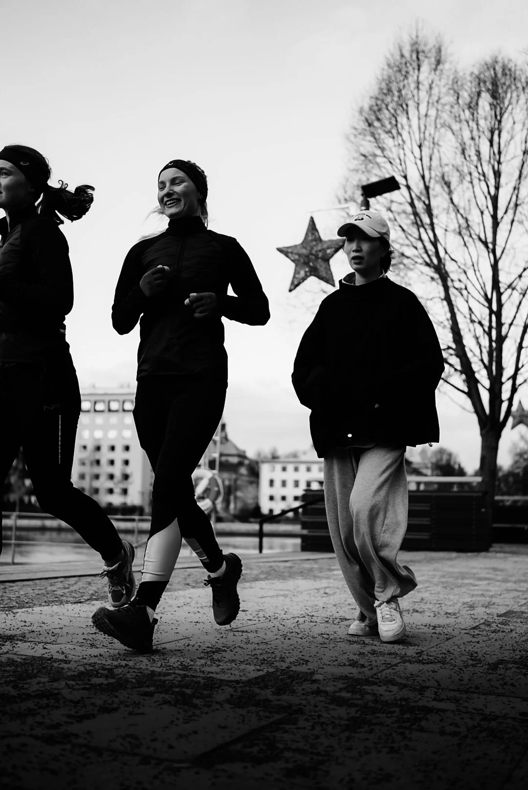 Three women and one girl jogging outdoors on a cloudy day, with leafless trees in the background and a large star decoration hanging in the distance.