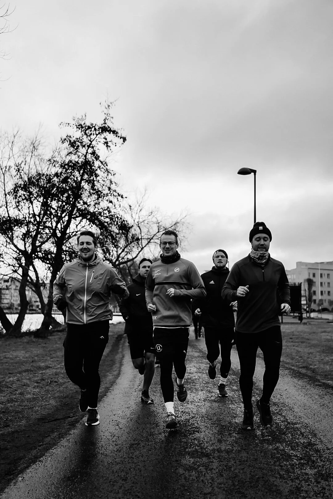 Group of five men running outdoors on a wet path with leafless trees and buildings in the background, overcast sky, black and white photo.