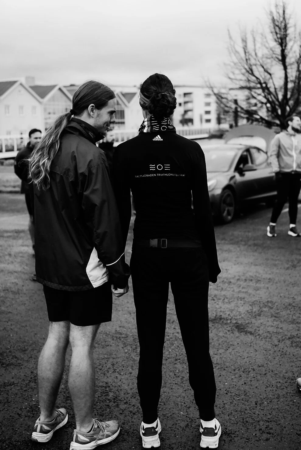 Two women in athletic clothing stand outdoors, talking to each other, with a group of people and buildings in the background.