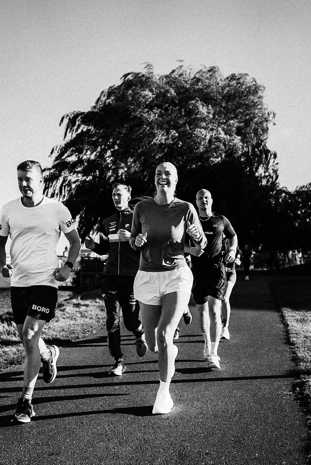A group of five people running outdoors on a paved path, smiling and appearing joyful, with a large tree and clear sky in the background.