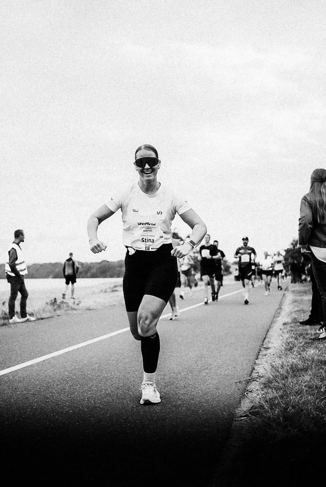 A woman with a big smile running in a marathon, wearing dark sunglasses, a white shirt, black shorts, and a marathon bib, with other runners and spectators in the background on a paved road.