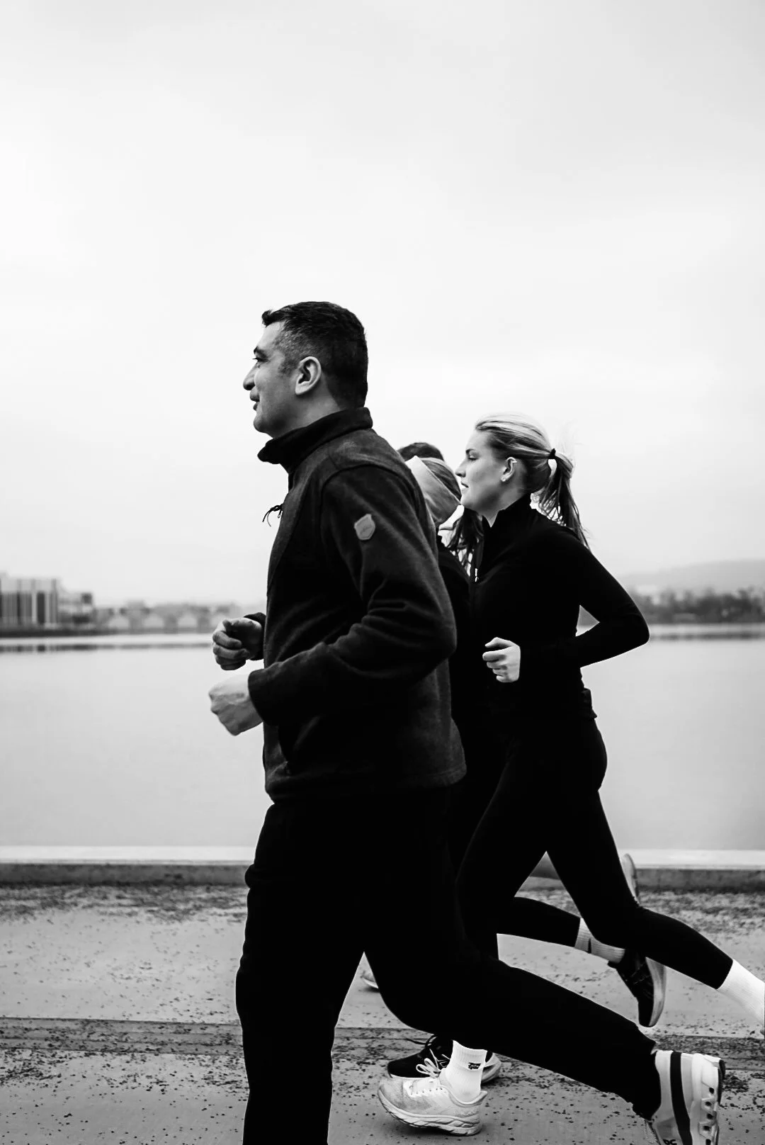 Black and white photo of three people jogging outdoors near a body of water, wearing athletic clothing.