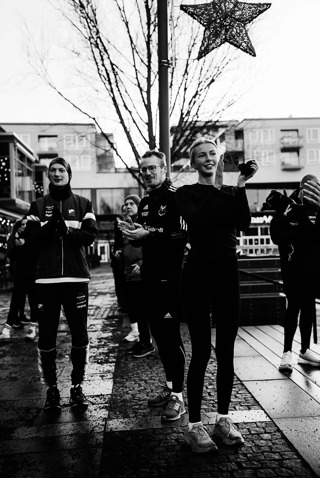Black and white photo of people in athletic gear gathered outdoors, some clapping, in front of a decorated lamppost with a star and streamers, with leafless trees and buildings in the background.