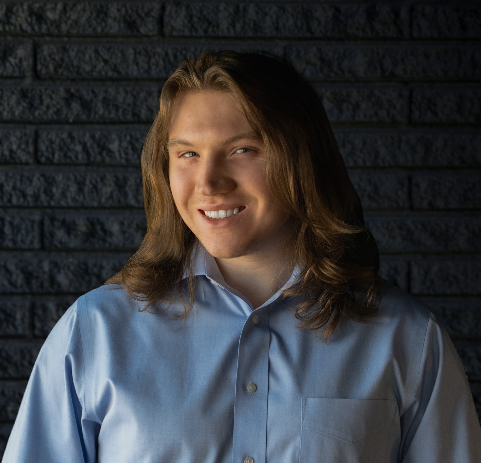 A young man with long brown hair, wearing a light blue dress shirt, smiling against a black background.