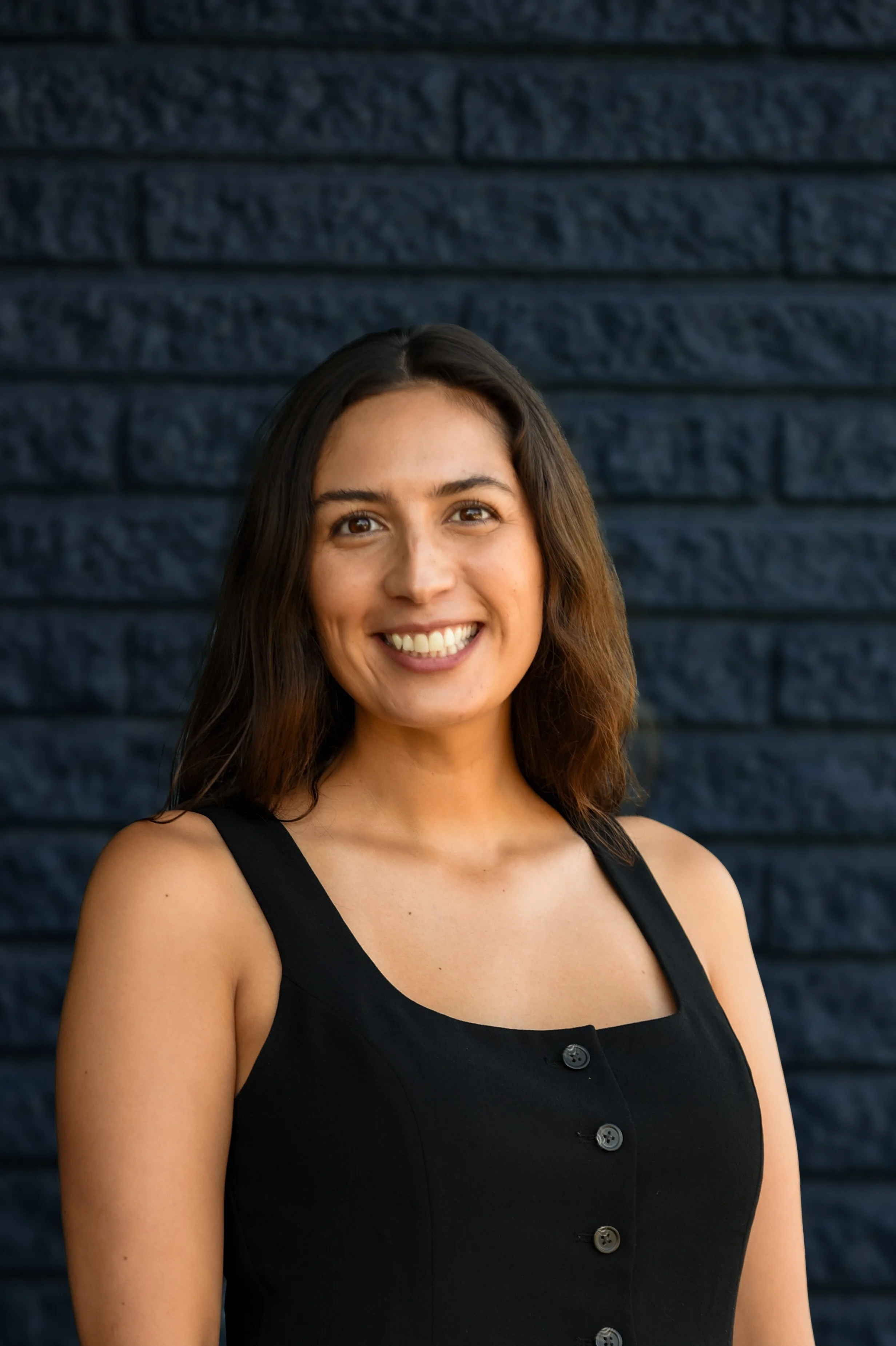 A woman with long brown hair smiling at the camera, wearing a sleeveless maroon top with buttons, against a black background.