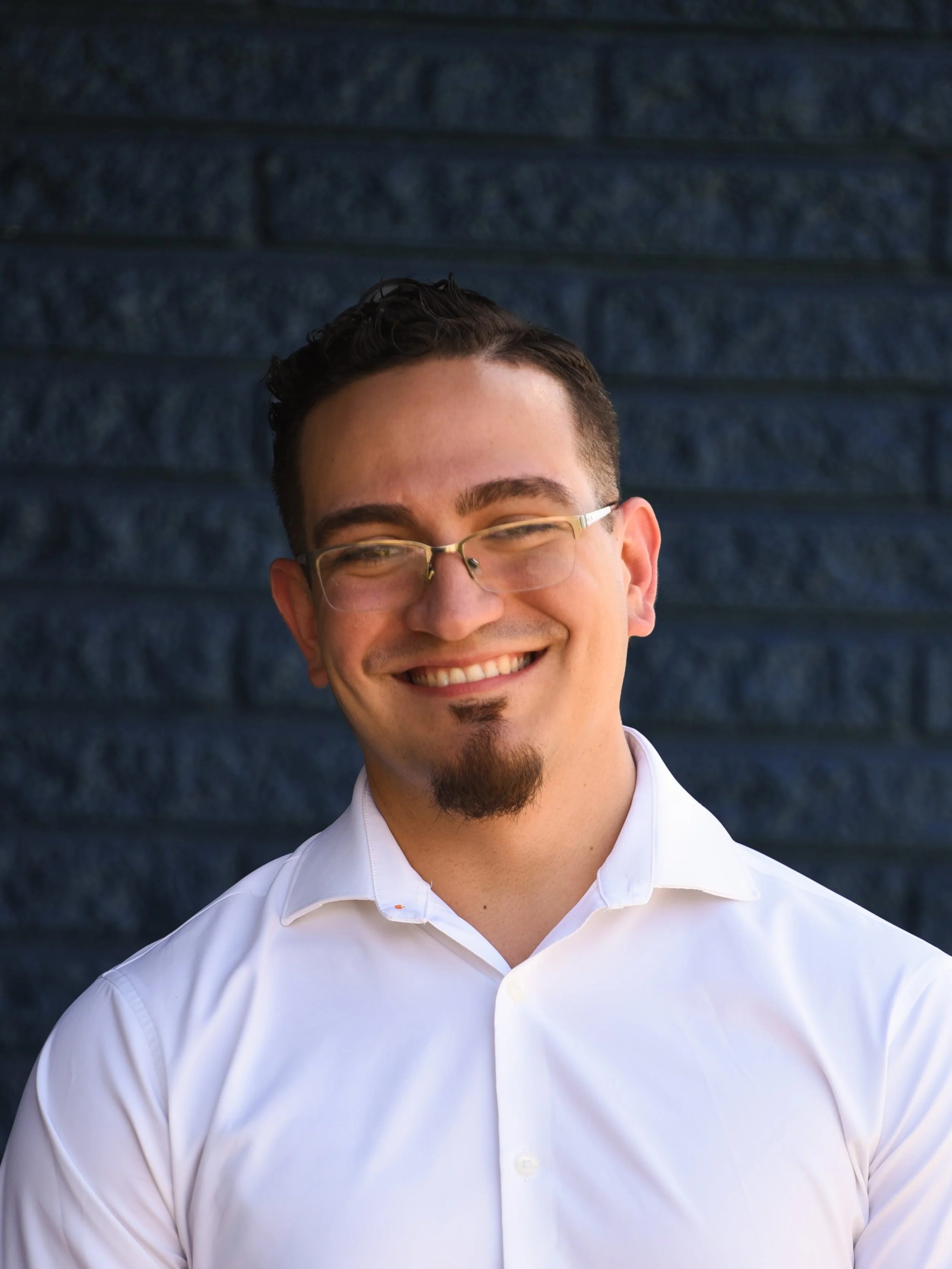 A young man with glasses and a goatee, smiling, wearing a light beige shirt, against a black background.