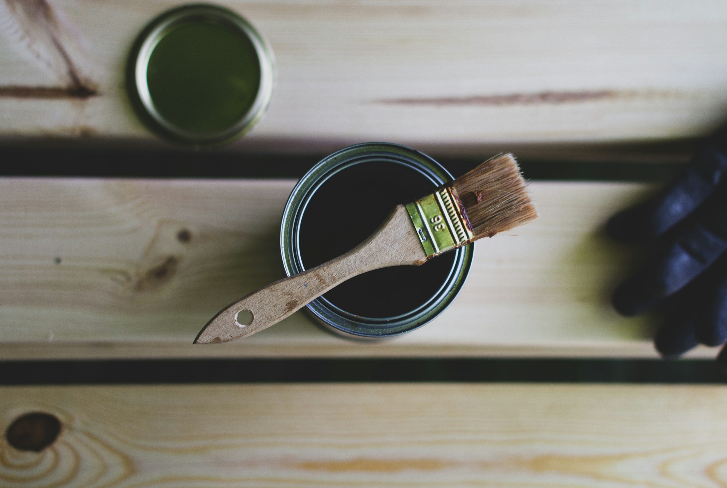 An open can of dark green paint with a paintbrush resting on the rim, placed on a light wooden surface, with another can of green paint and a pair of black gloves nearby.