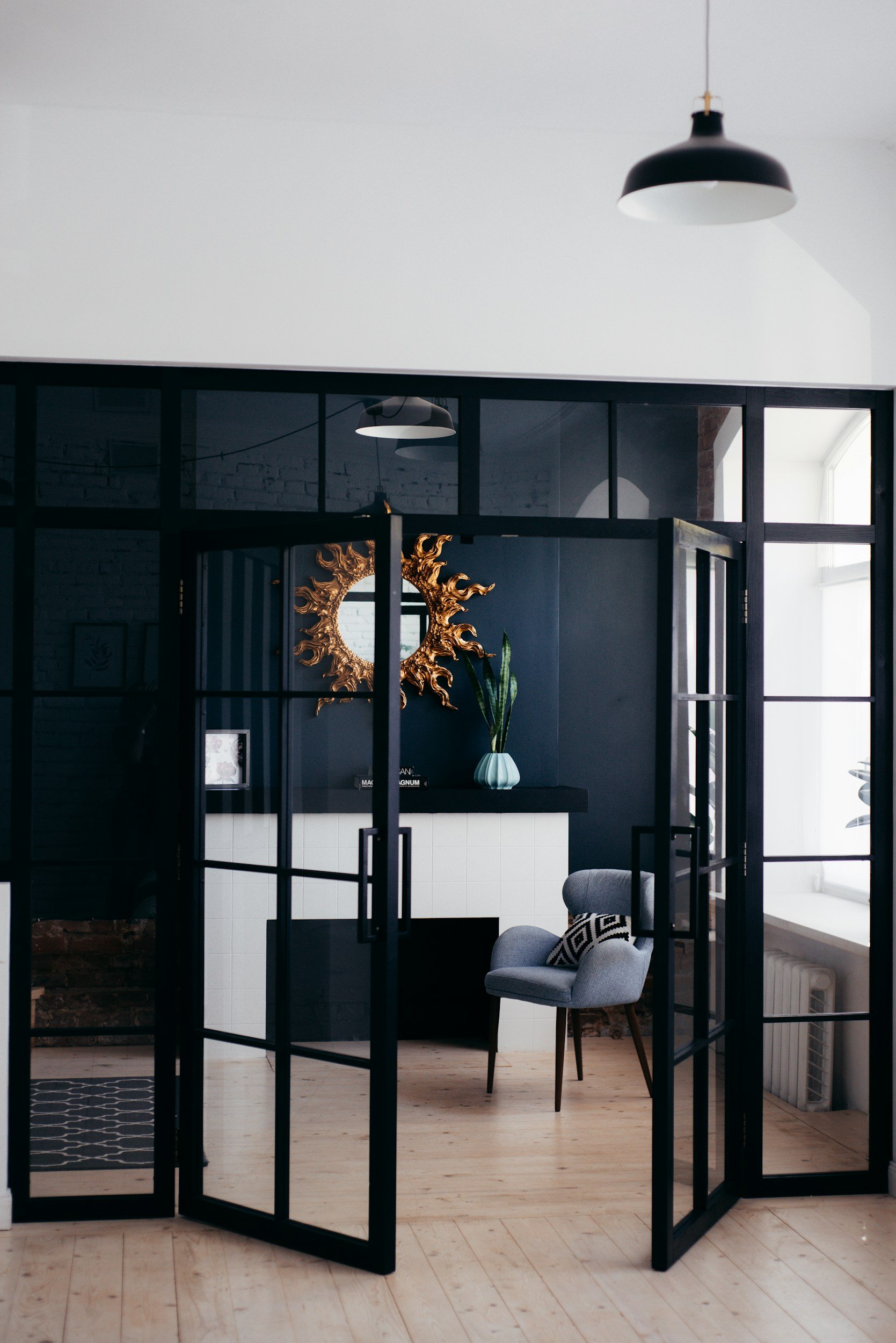 Modern interior space with black-framed glass doors, a gray armchair with a patterned pillow, a black shelf with a tall plant and decorative sun mirror, and wooden flooring with natural light from large windows.