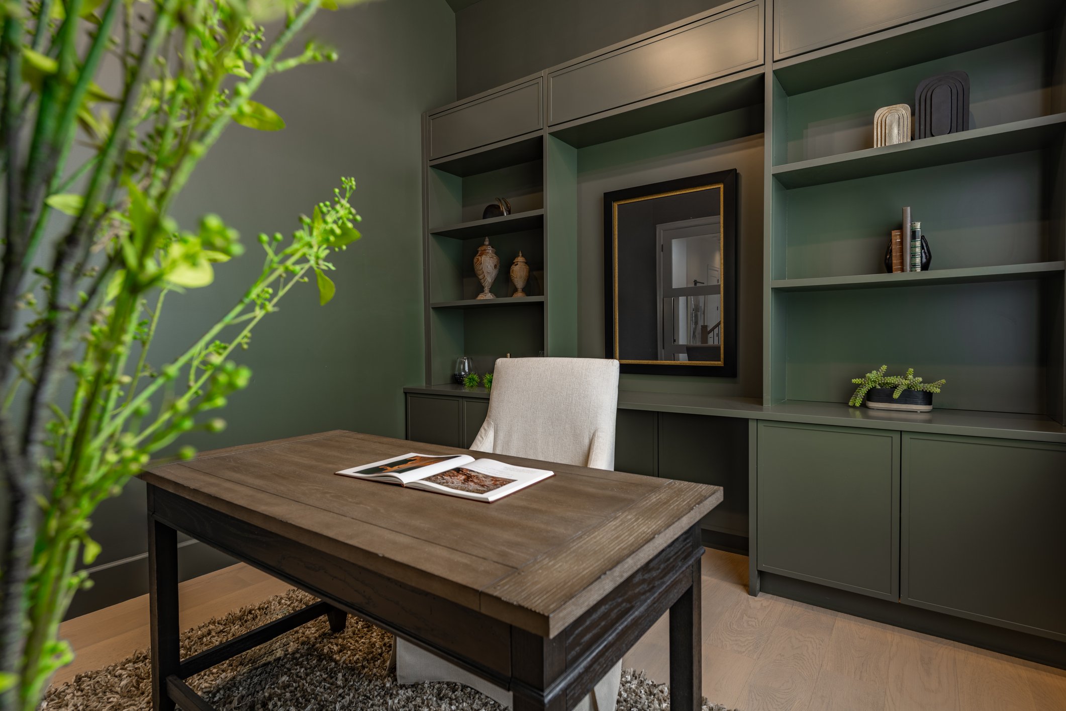 Home office with green walls, a wooden desk, white upholstered chair, and built-in green bookshelf with decorative items and books, a mirror, and a small plant.