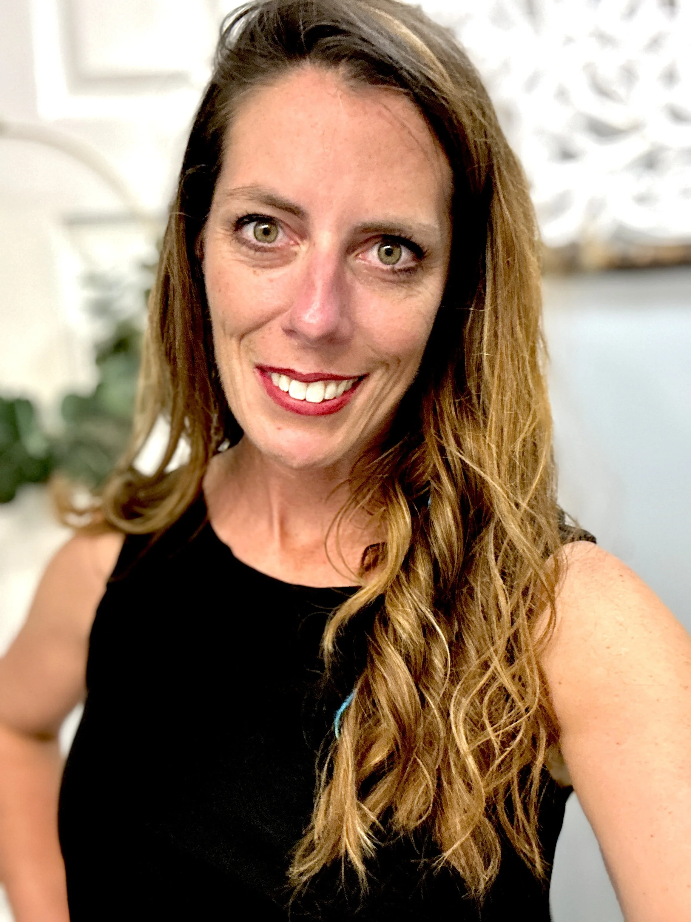 A woman with long, wavy, reddish-brown hair, green eyes, and red lipstick smiling at the camera, wearing a black sleeveless top indoors.