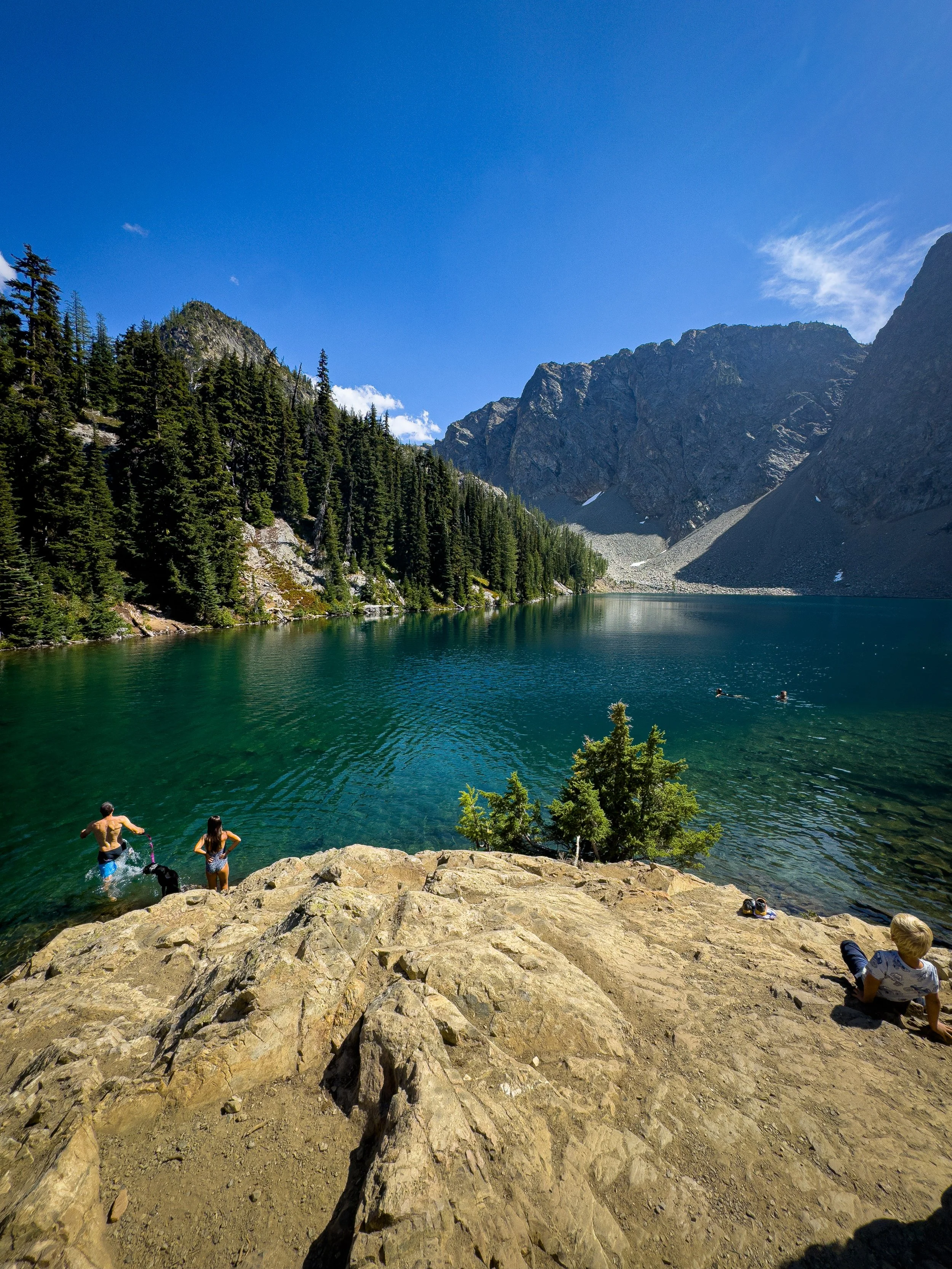 People swimming and resting on a rocky shoreline beside a clear alpine lake beneath steep peaks.