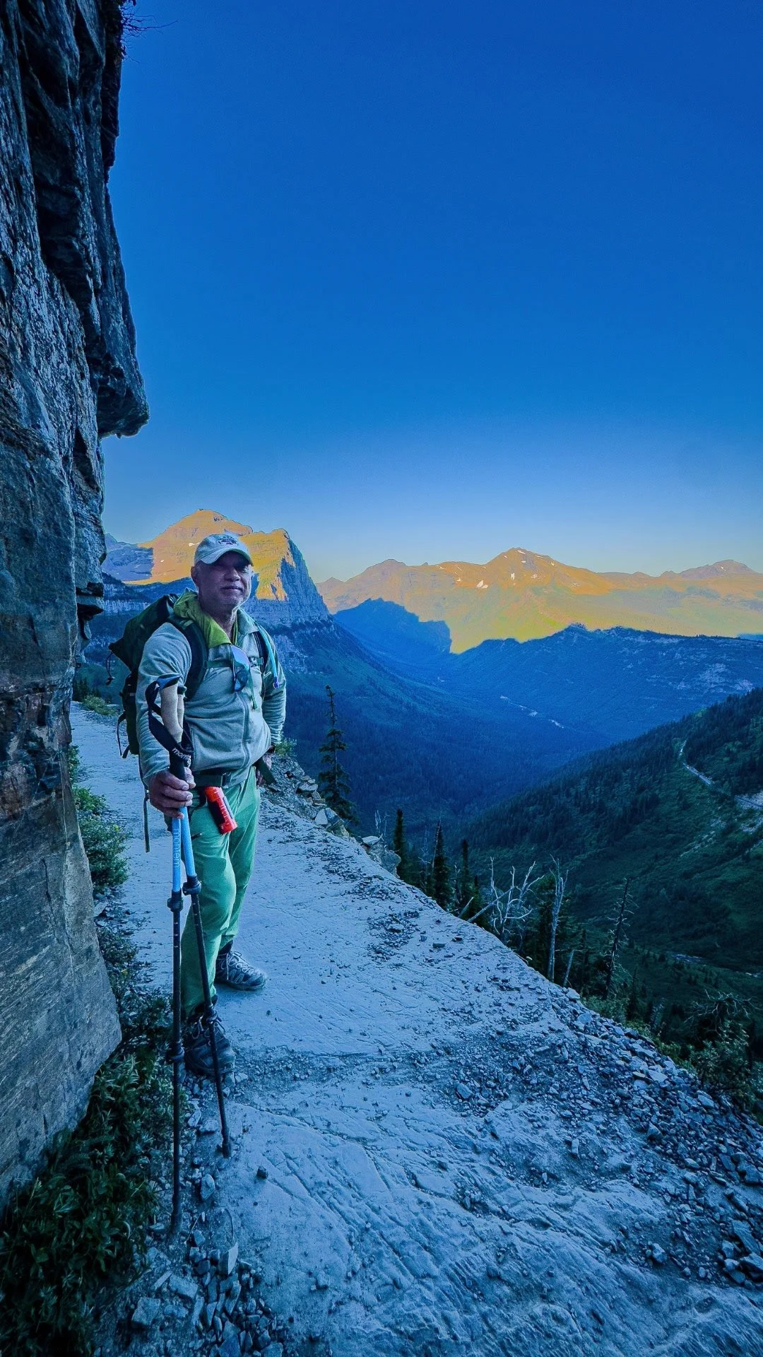 Sundar with hiking poles on the narrow Highline Trail beside a rock wall, above a forested valley and distant mountains.