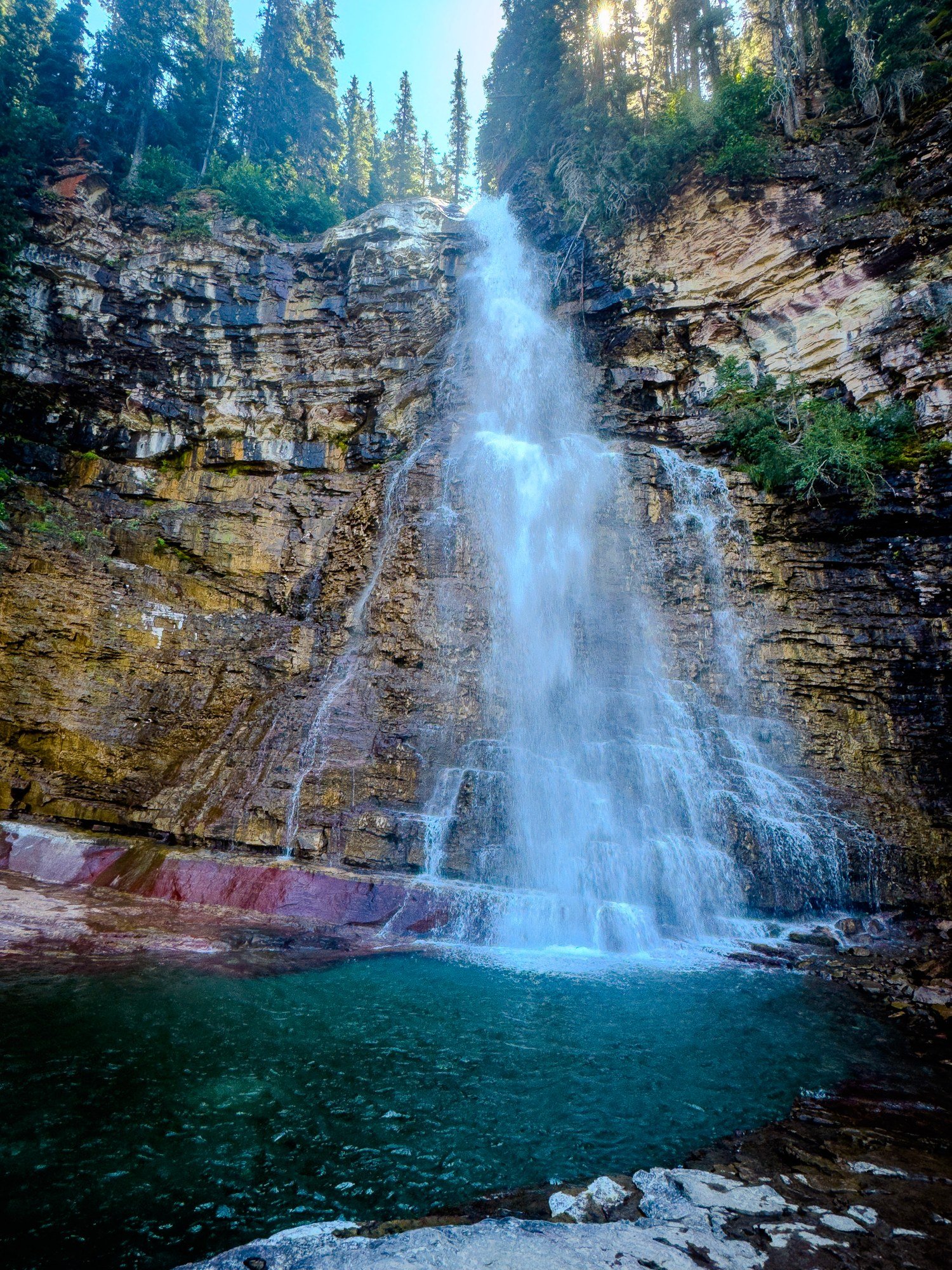 A waterfall cascades over layered rock into a turquoise pool beneath a forested slope.