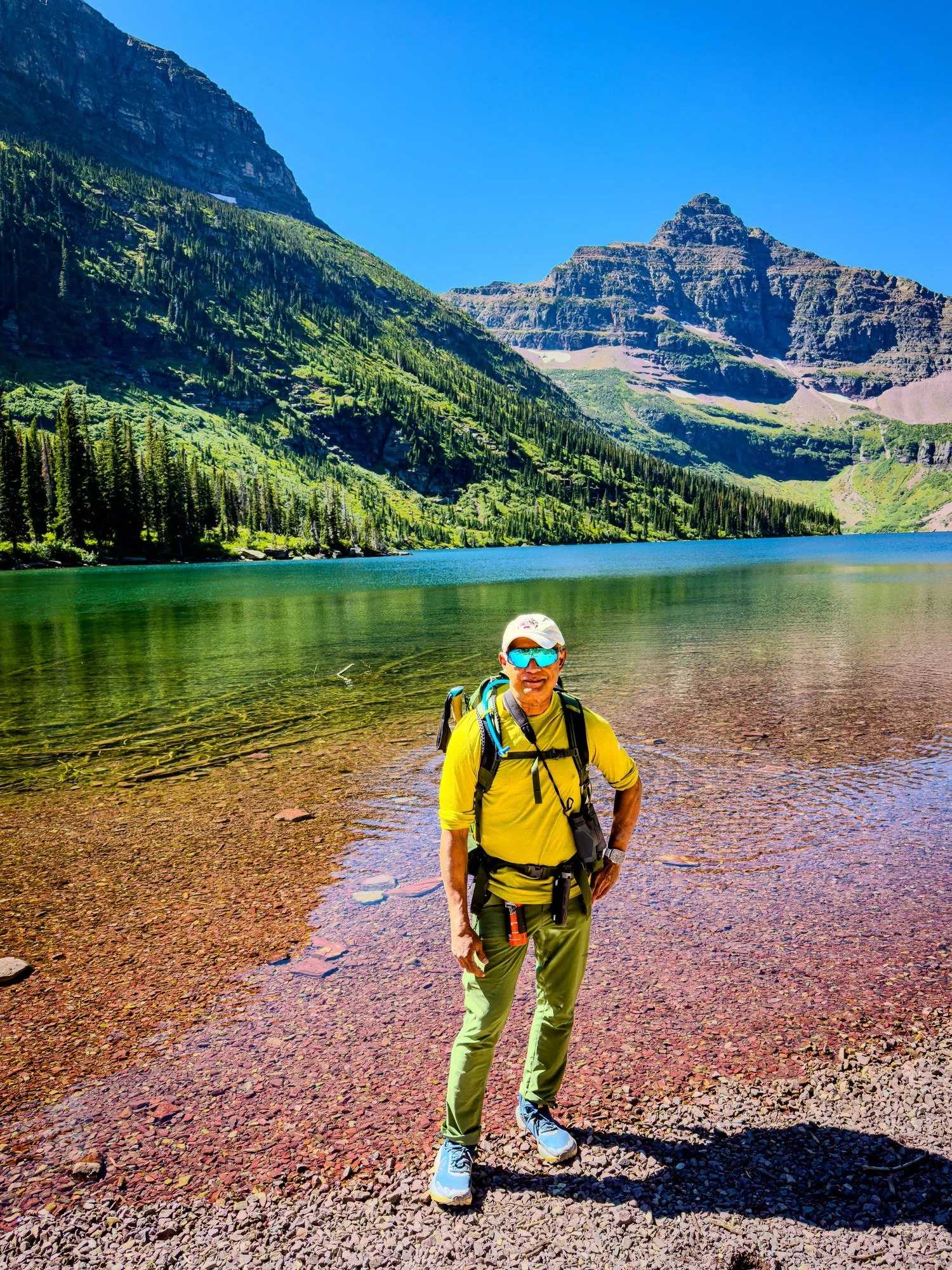 Sundar in hiking gear beside a clear mountain lake, with forested slopes and rocky peaks behind him.