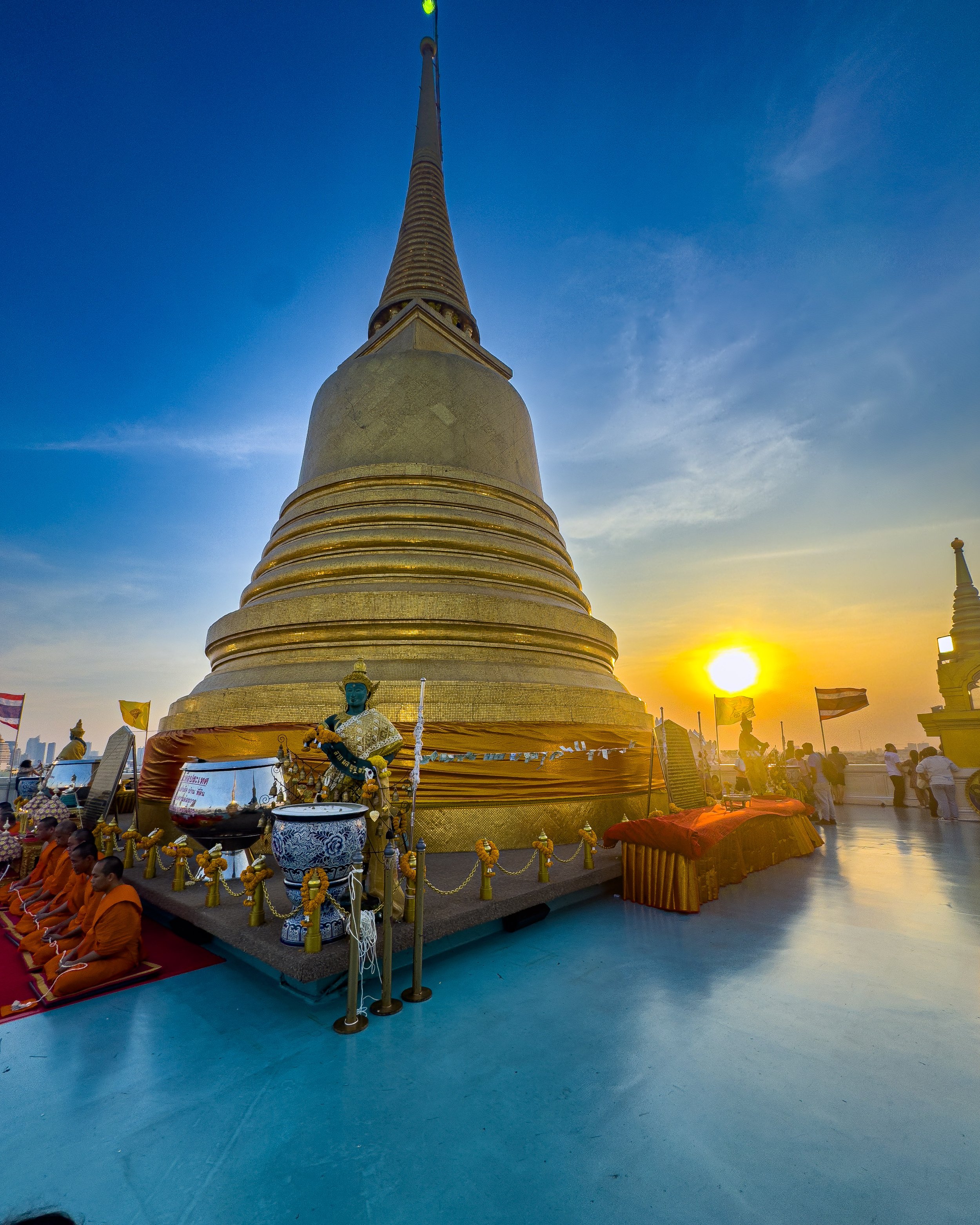 Golden stupa in Thailand at sunset, with orange-robed monks seated at lower left and the setting sun glowing on the right.
