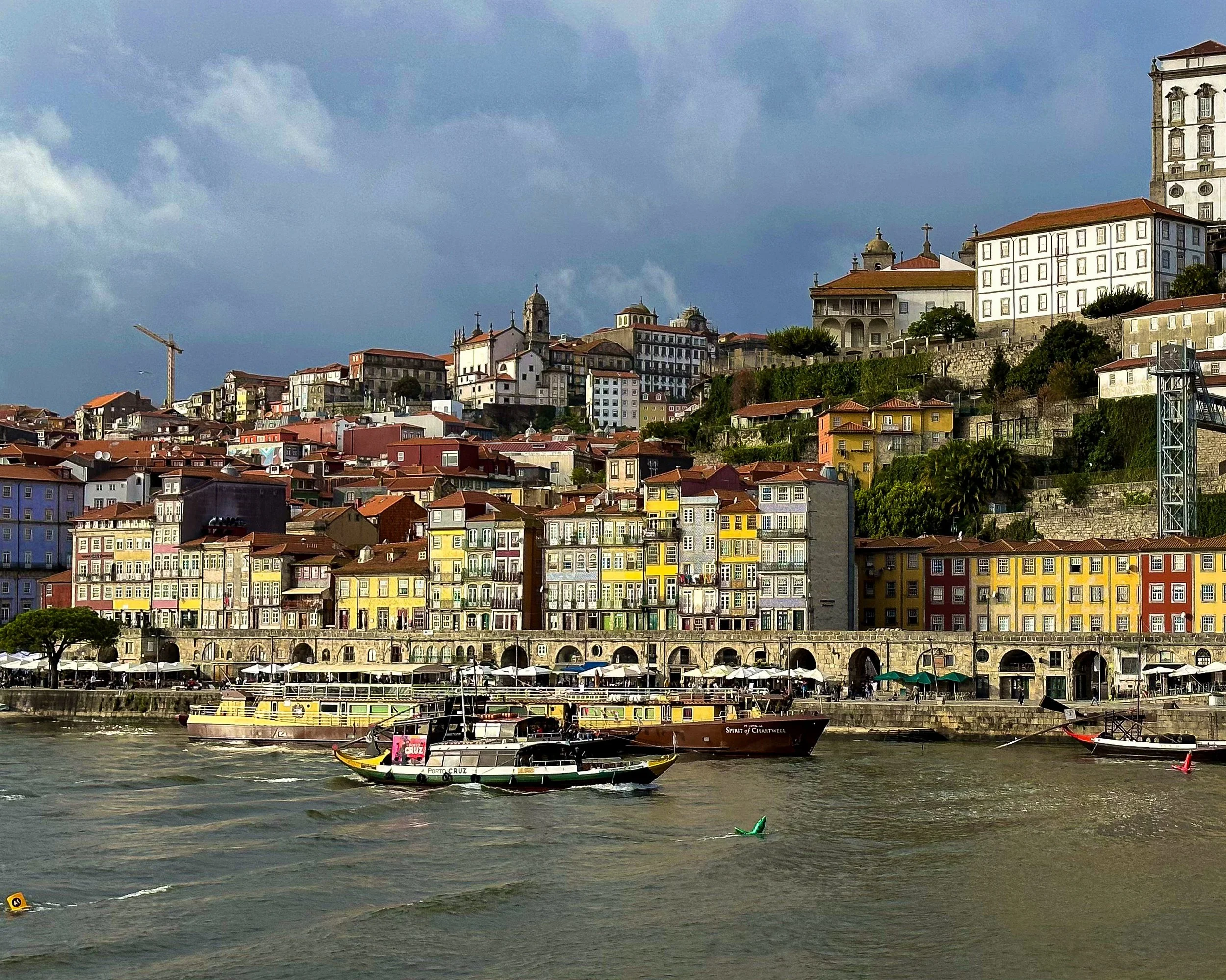 Colorful Ribeira waterfront buildings in Porto, Portugal, with boats on the Douro River beneath a cloudy sky.