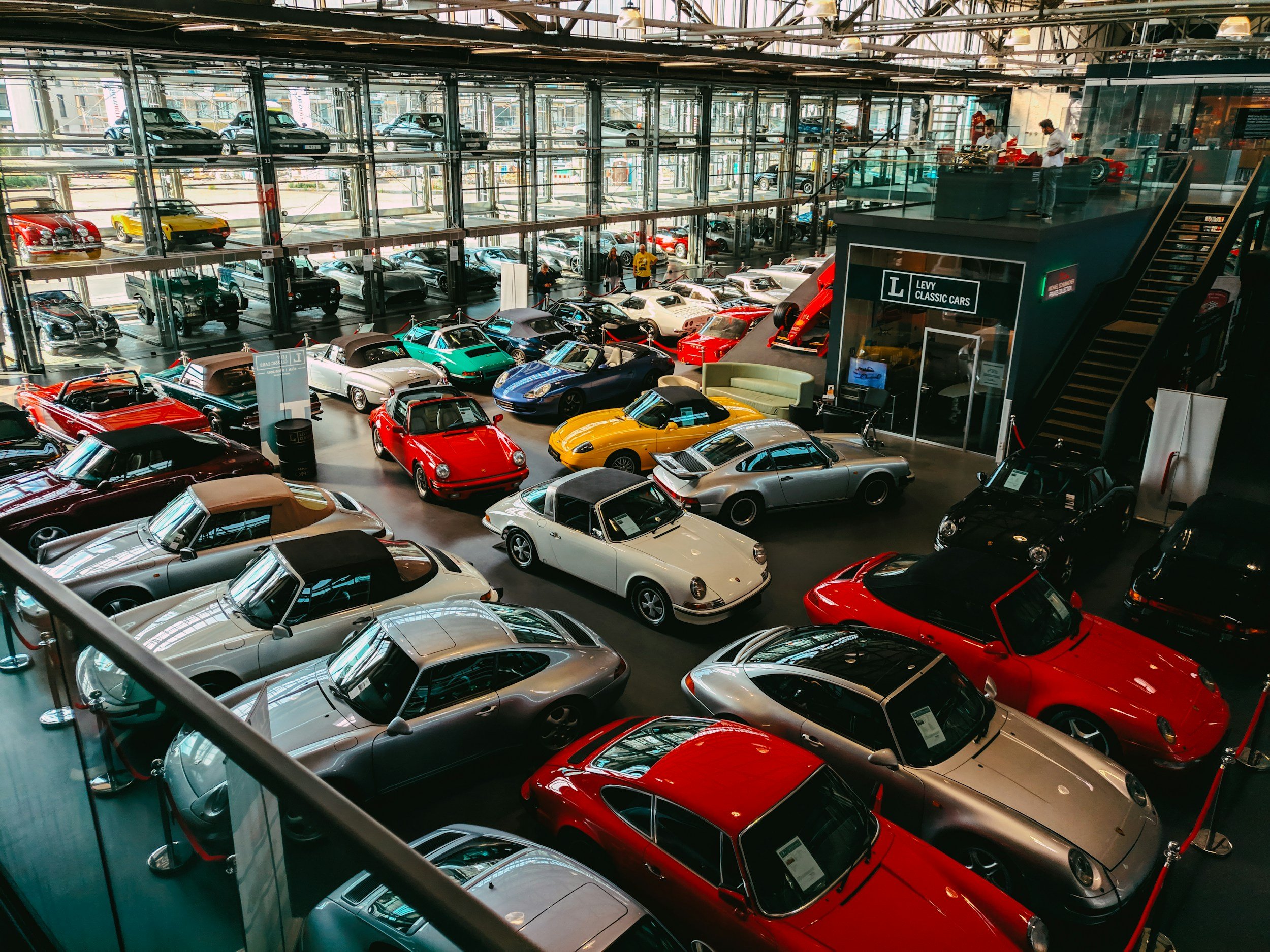 Collection of classic cars displayed in a showroom with glass walls and multiple floors. Various models and colors are visible, including red, silver, and blue sports cars. A sign for "Levy Classic Cars" is present.