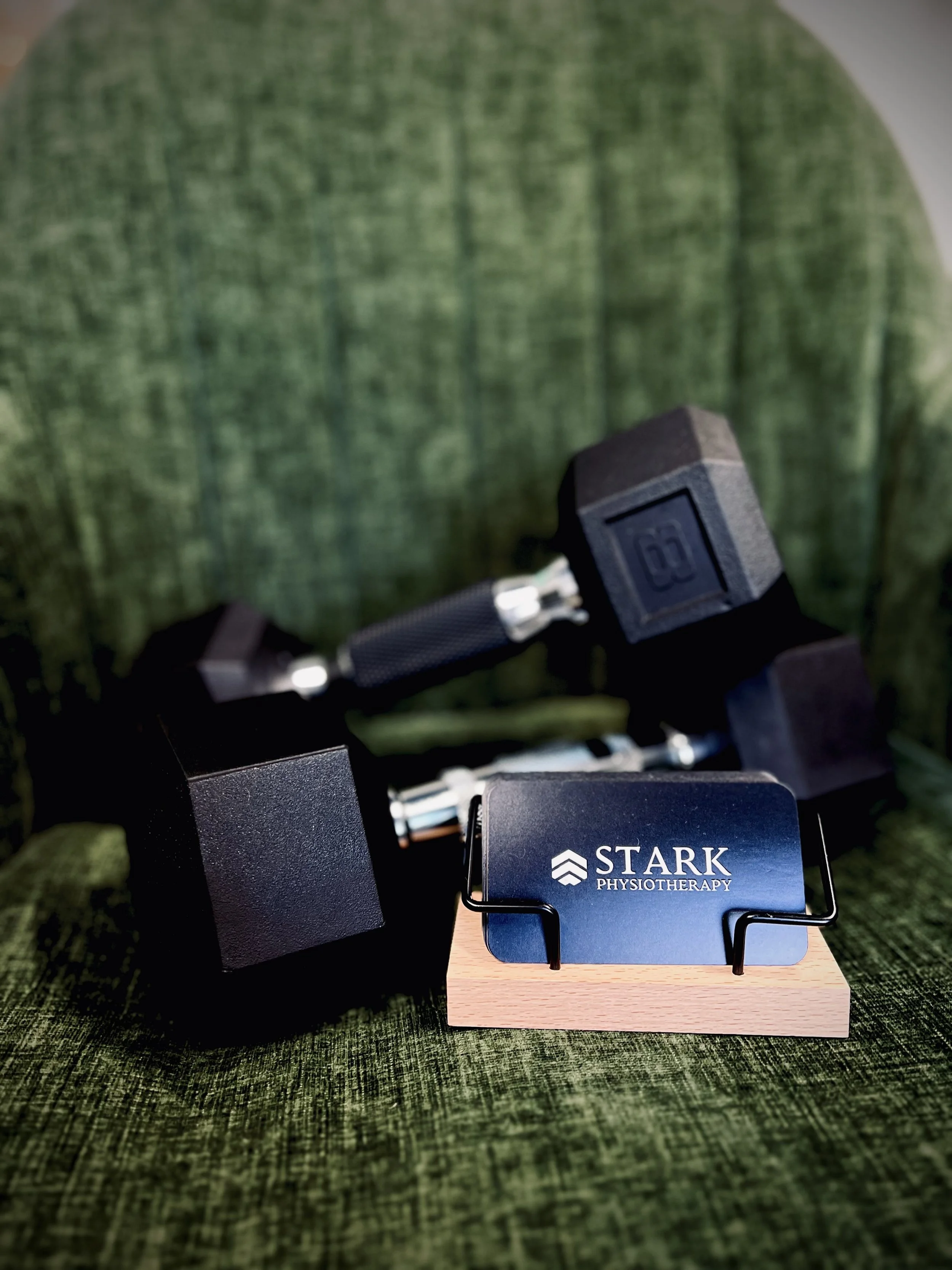 Dumbbells and a Stark Physiotherapy branded card stand on a green textured background, indicating a fitness or physiotherapy setting.